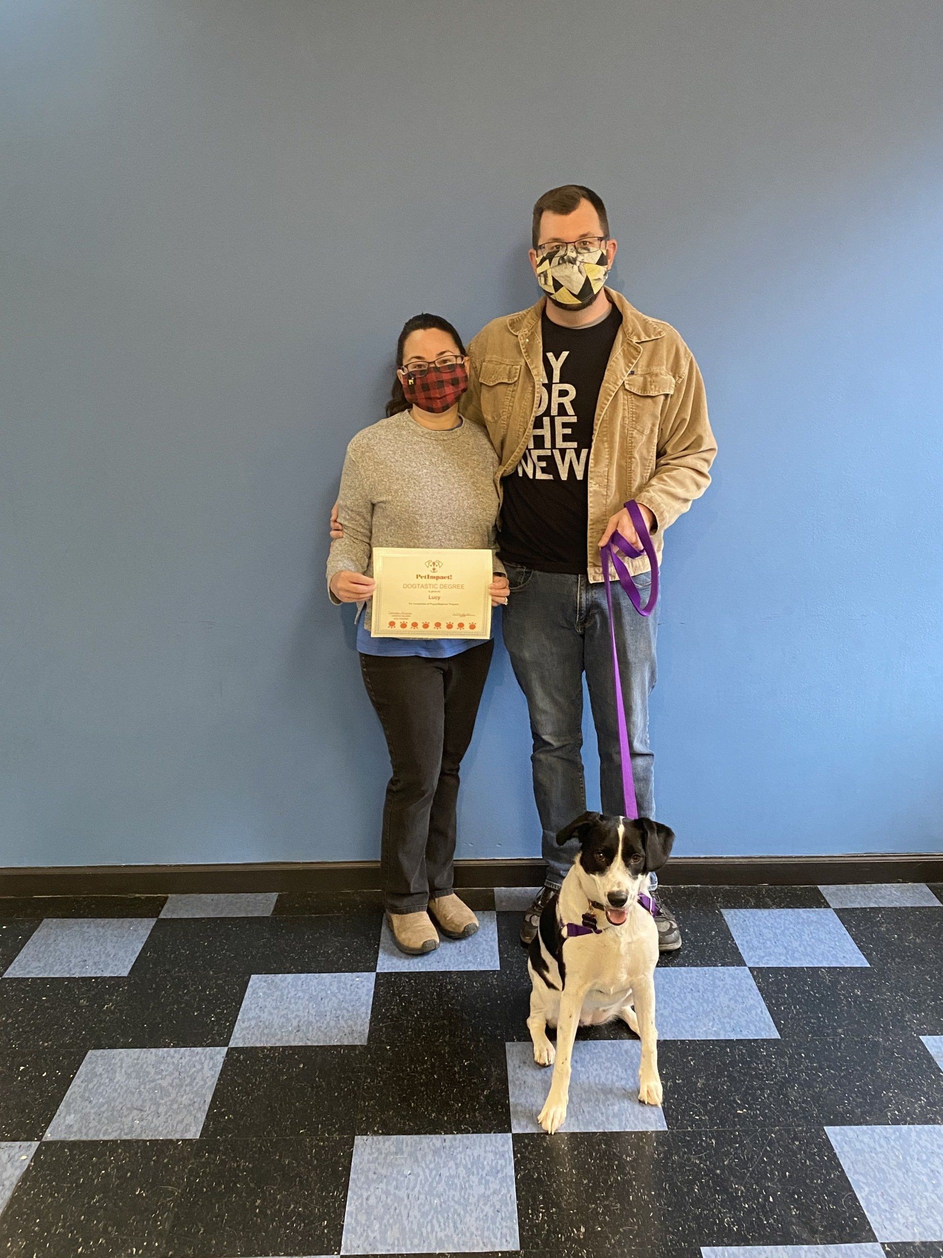 Couple and dog with certificate posing by blue wall. Dog is black and white. Everyone wearing masks.
