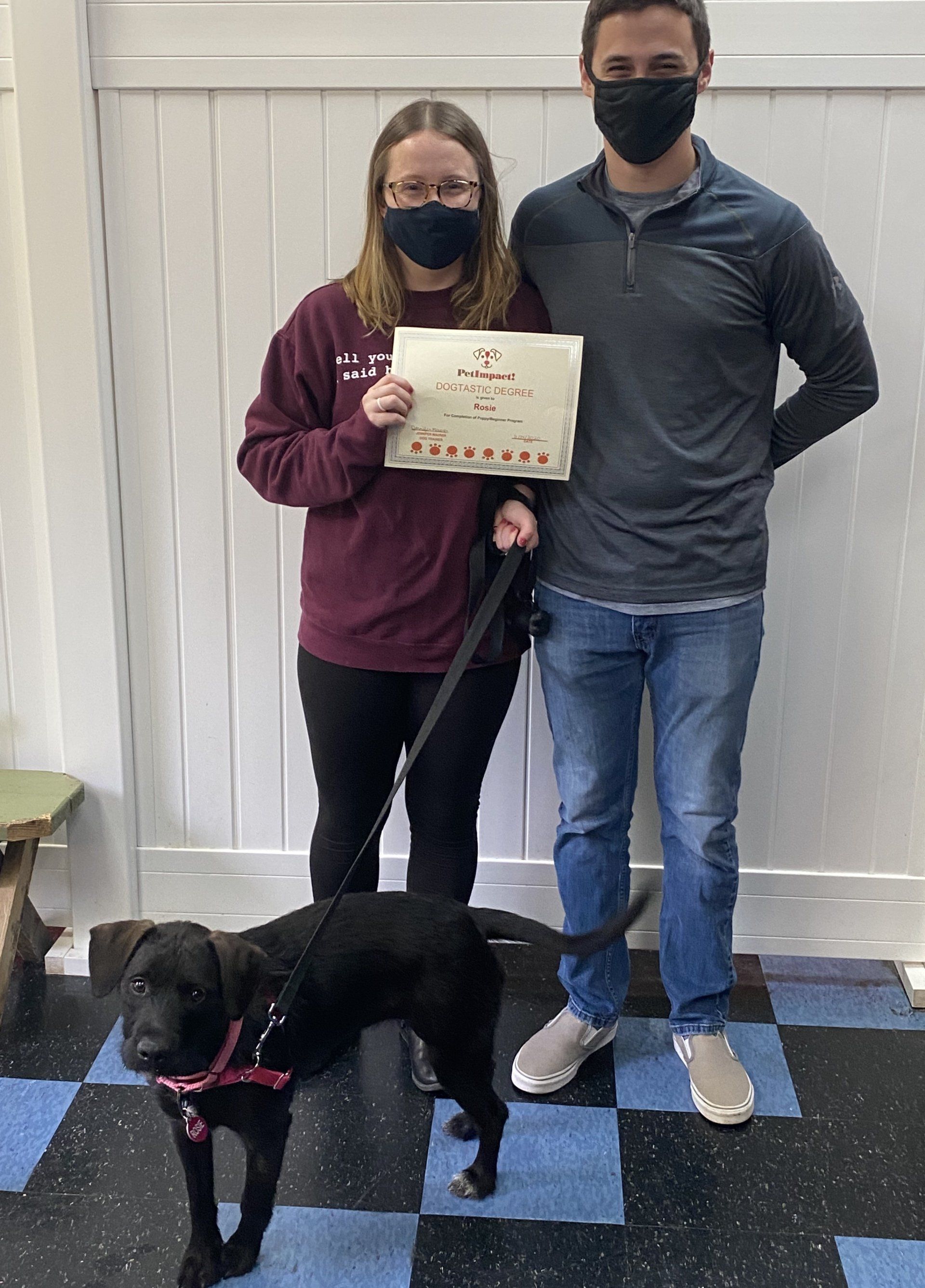 A couple and a black dog posing; woman holding a paper, all wearing masks, in a room.