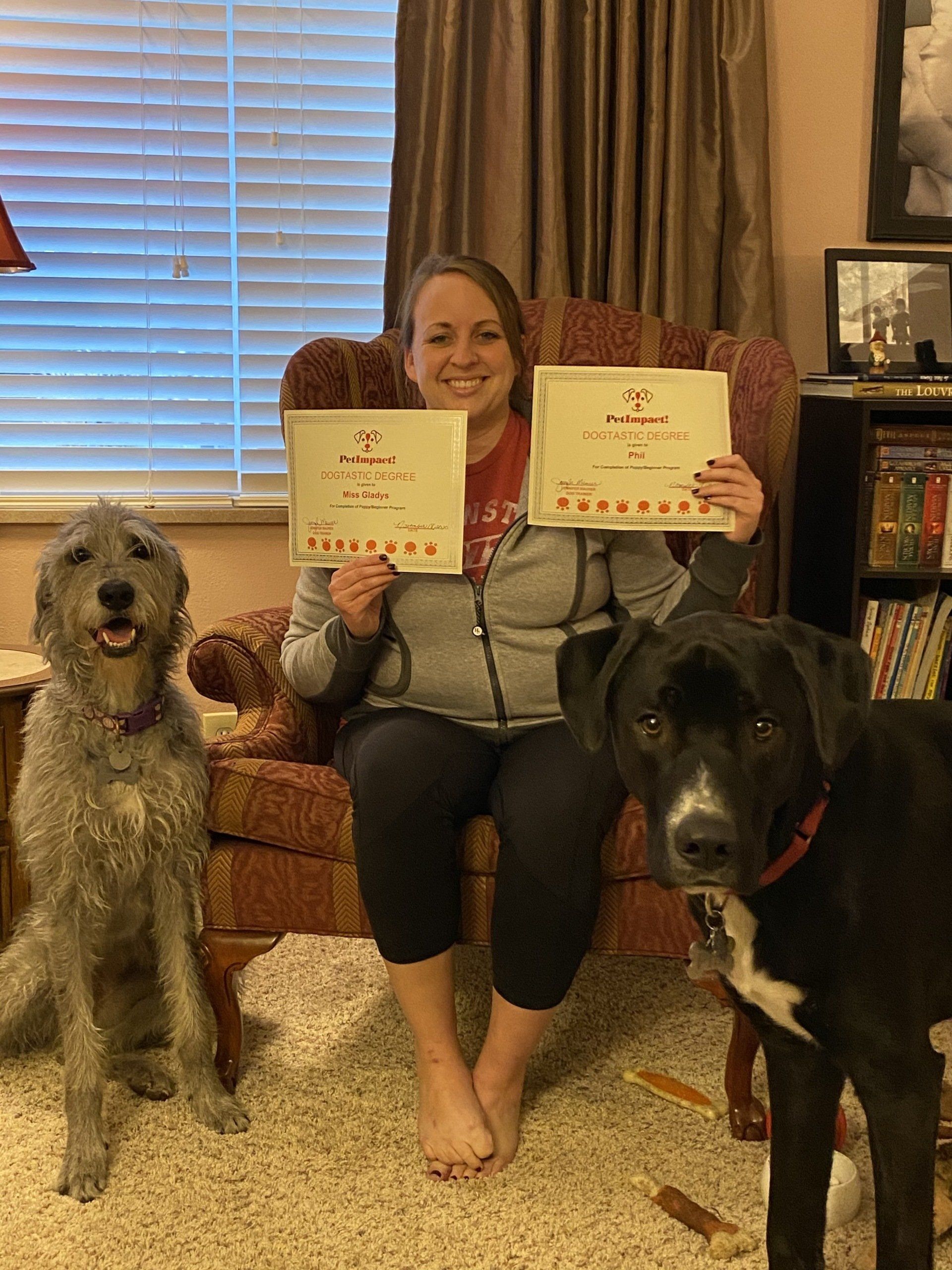 Woman sitting with two dogs holding award certificates, smiling. Indoors, warm tones.