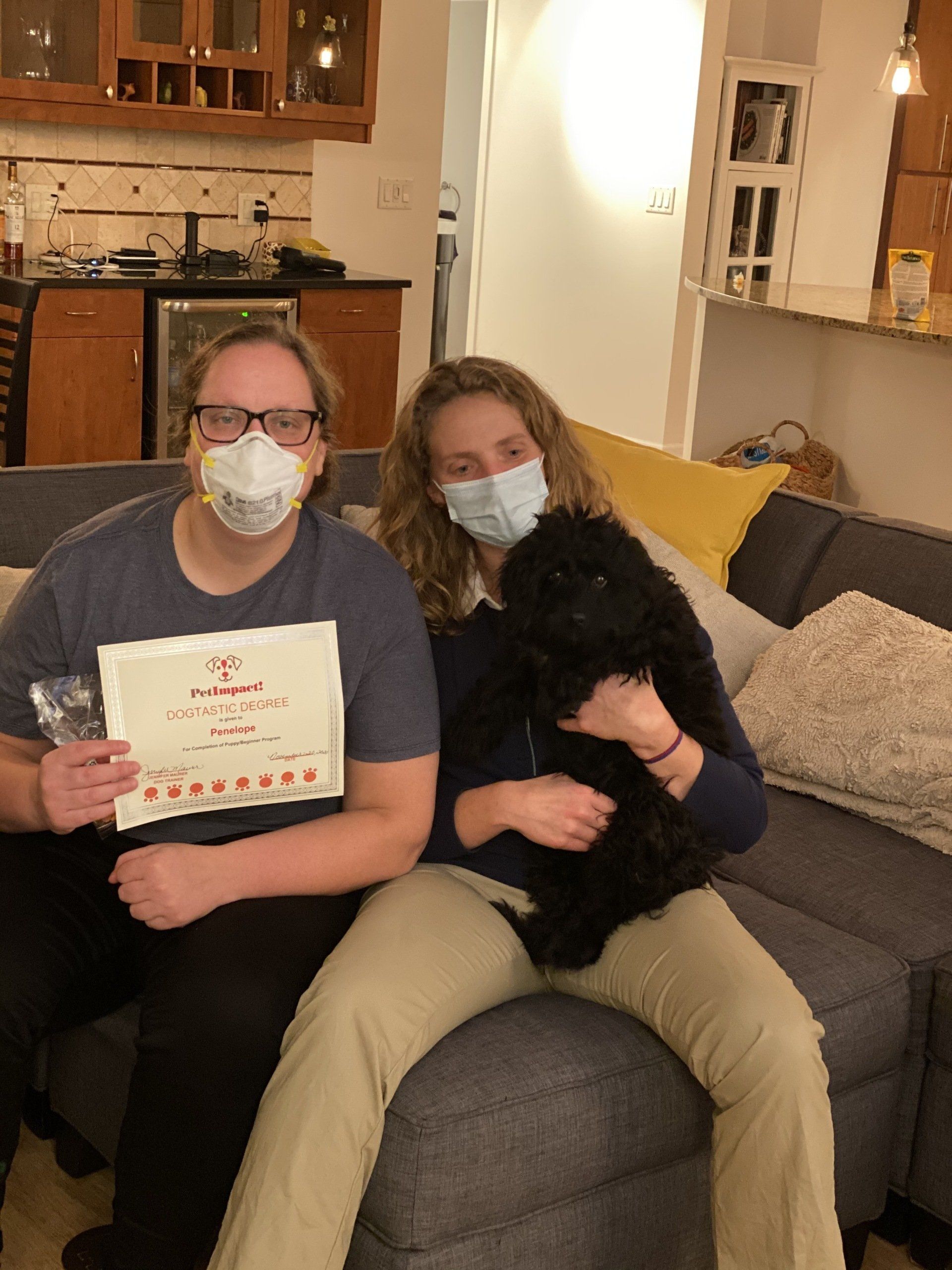 Two people wearing masks and a dog on a couch. One person holds a certificate and a drink.