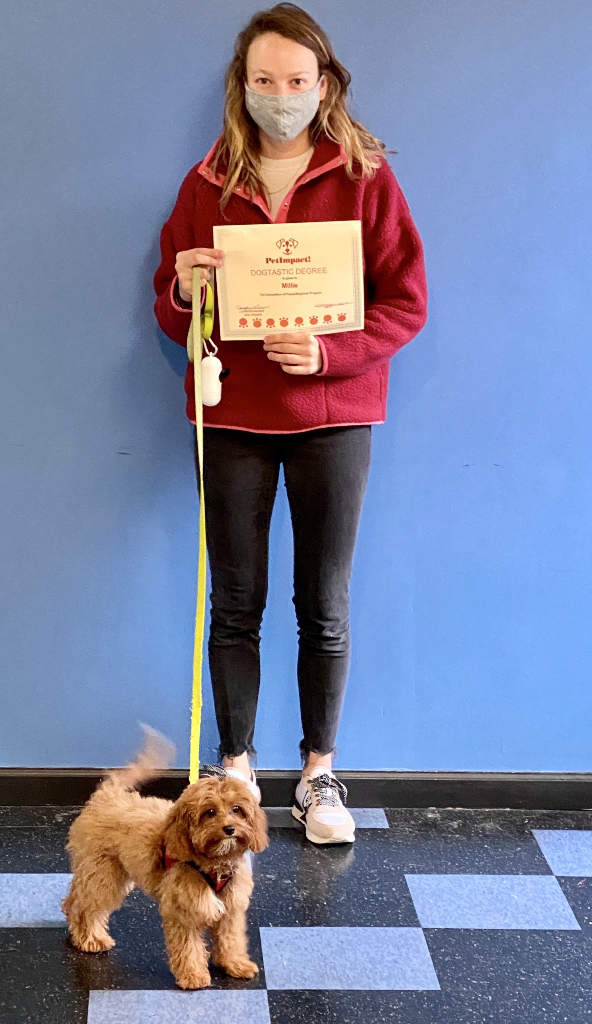 Woman in red jacket holds certificate, standing with a small, brown dog on leash by a blue wall.