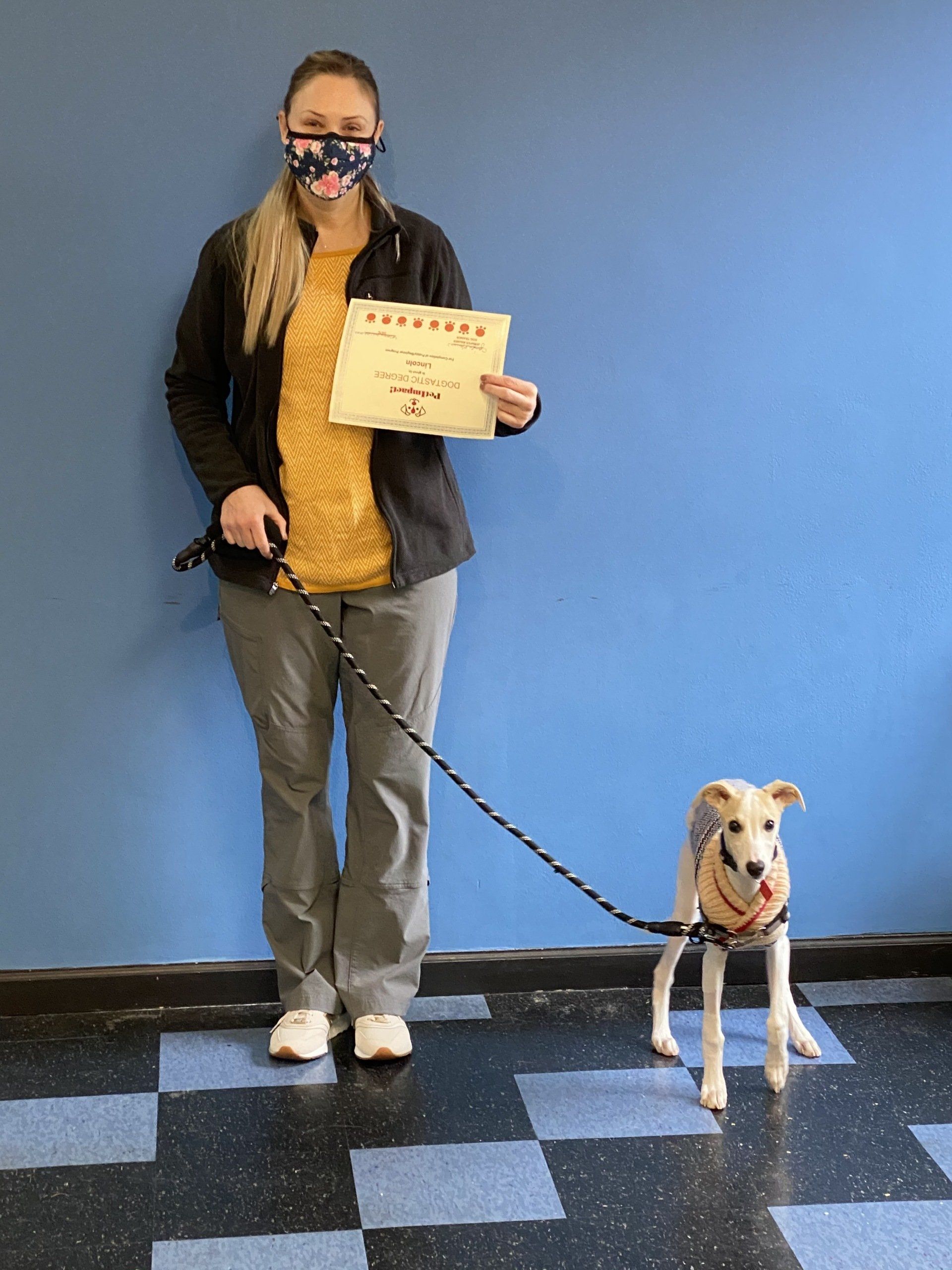 Woman in mask holding certificate, leash with dog in a vest, standing in front of a blue wall.