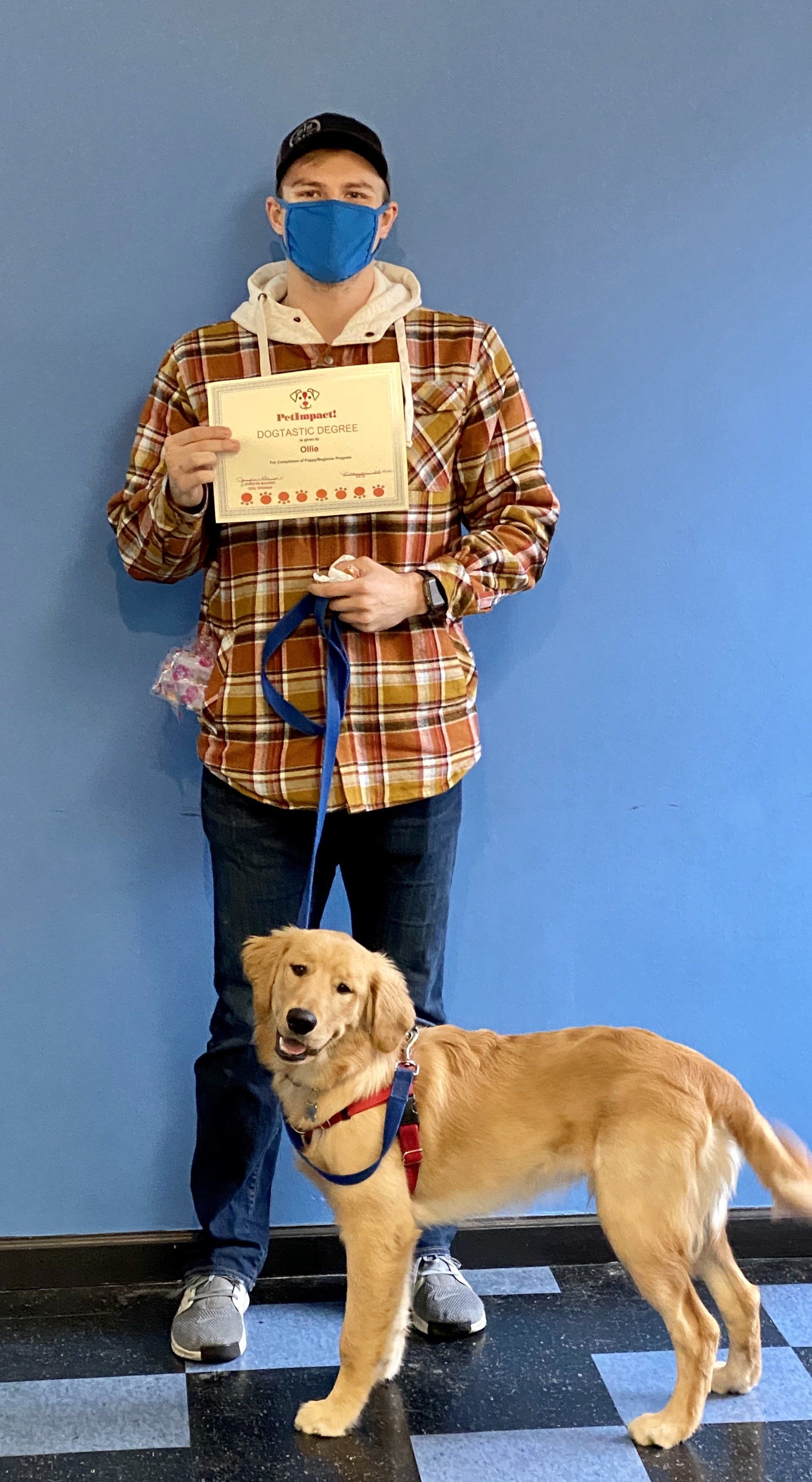 Man in plaid holding certificate, smiles with golden retriever; standing in front of blue wall.