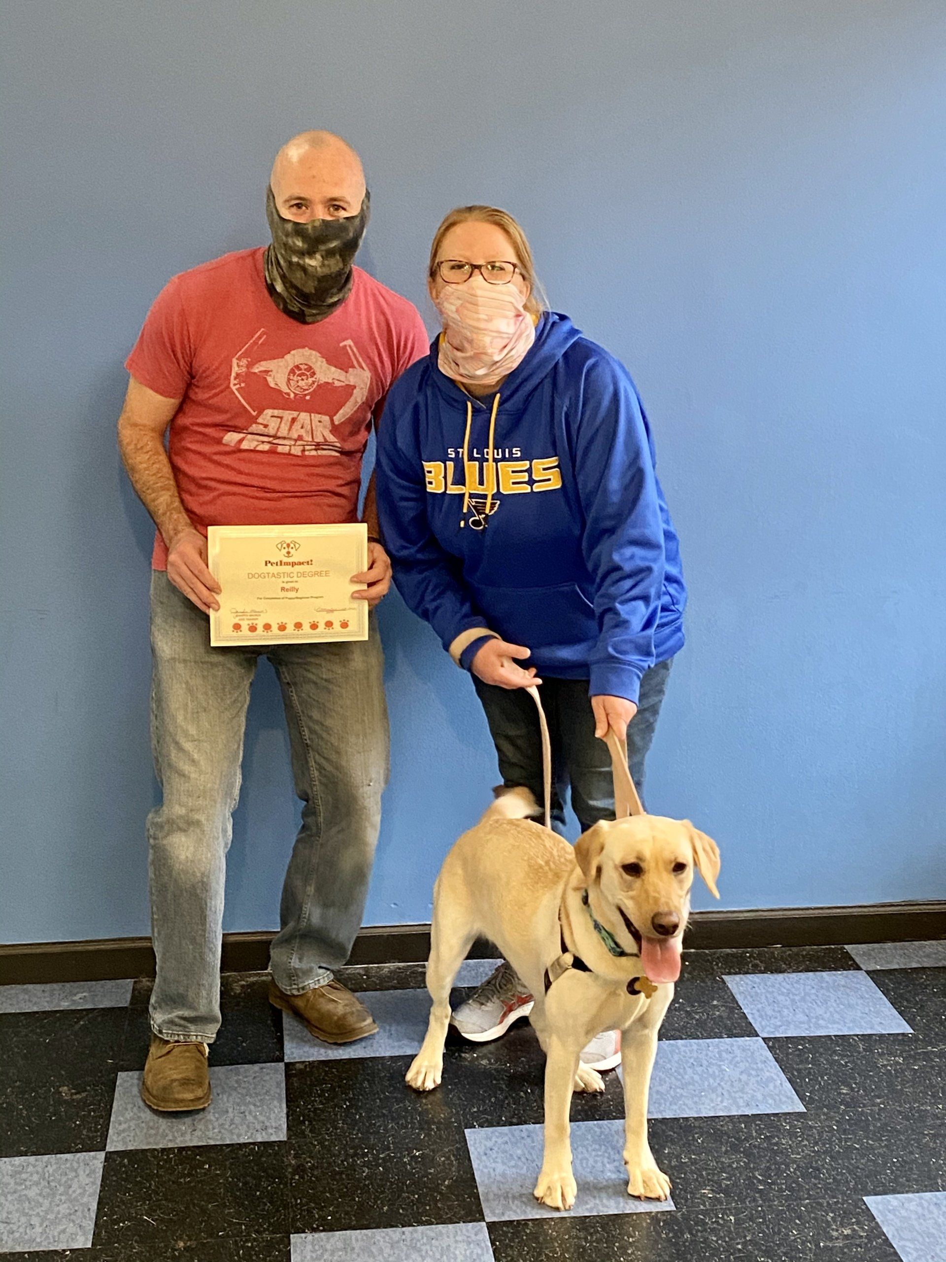 Two people with a yellow lab holding a certificate in front of a blue wall.