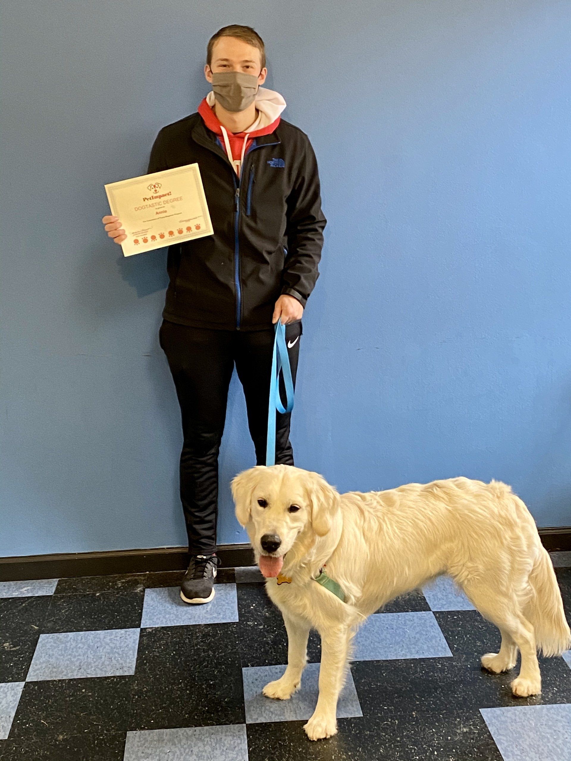 Man in a mask and dog with a leash stand near a blue wall and a checkered floor. The man holds a paper.