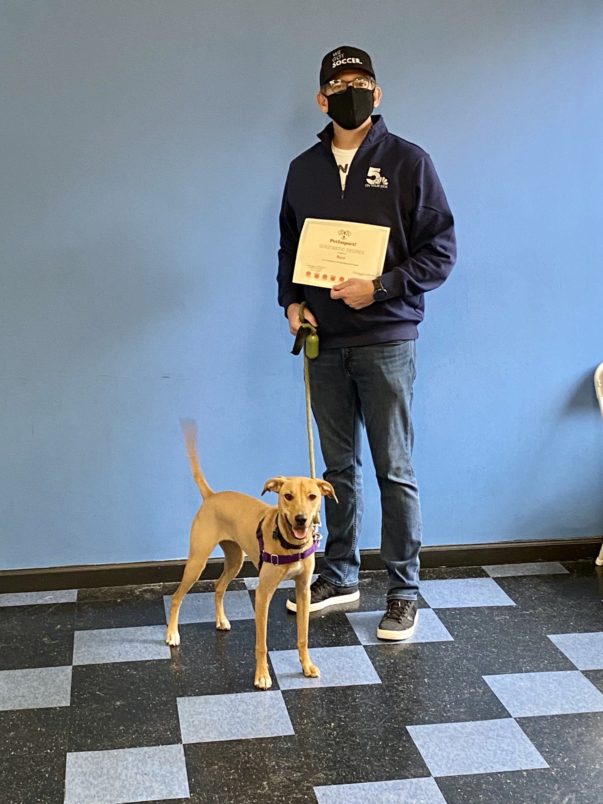 Man holding certificate stands with tan dog in front of blue wall. Checkered floor.