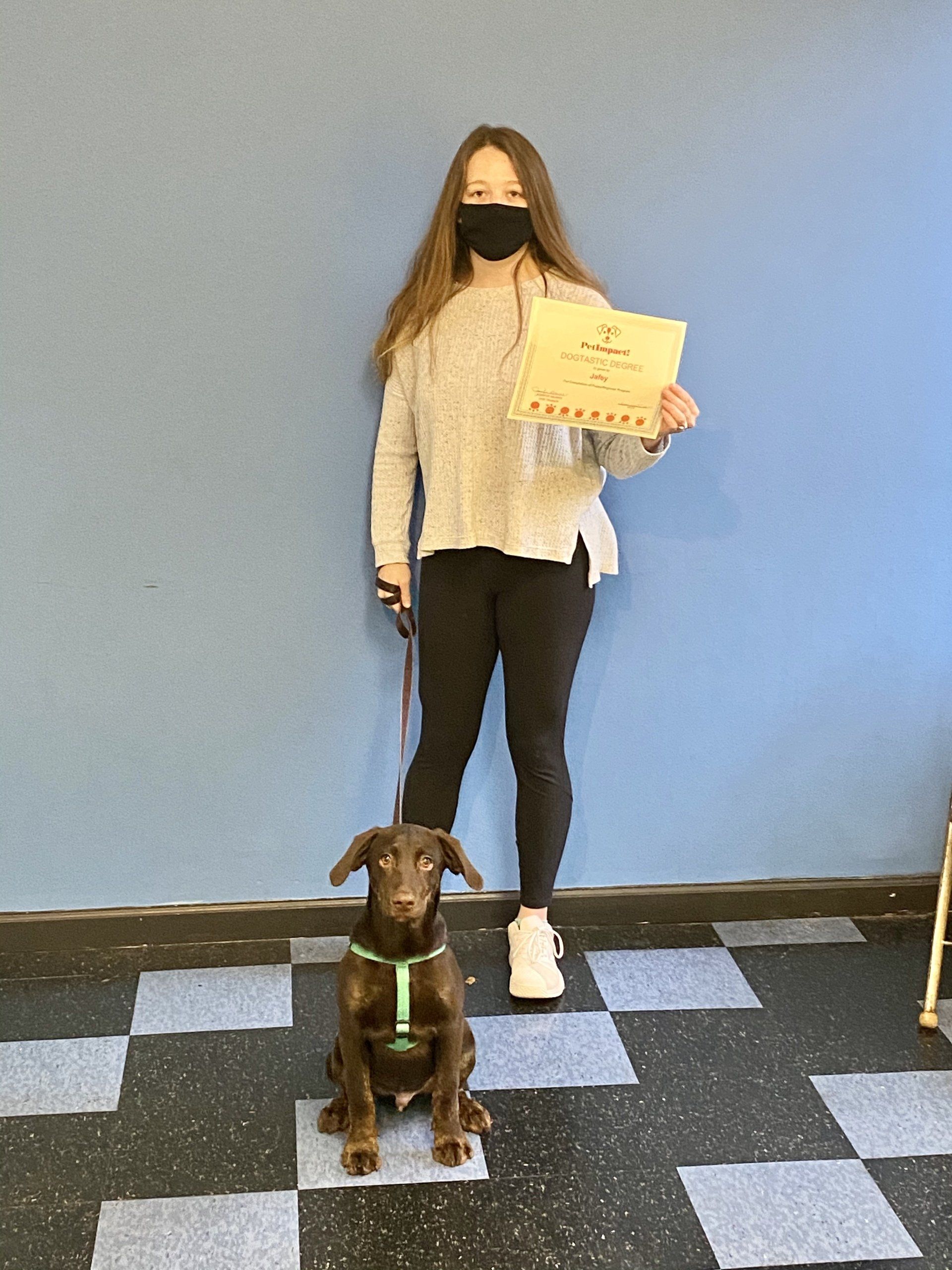 Woman with dog, holding certificate. Dog is brown and wearing a harness. Blue wall and checkered floor in the background.