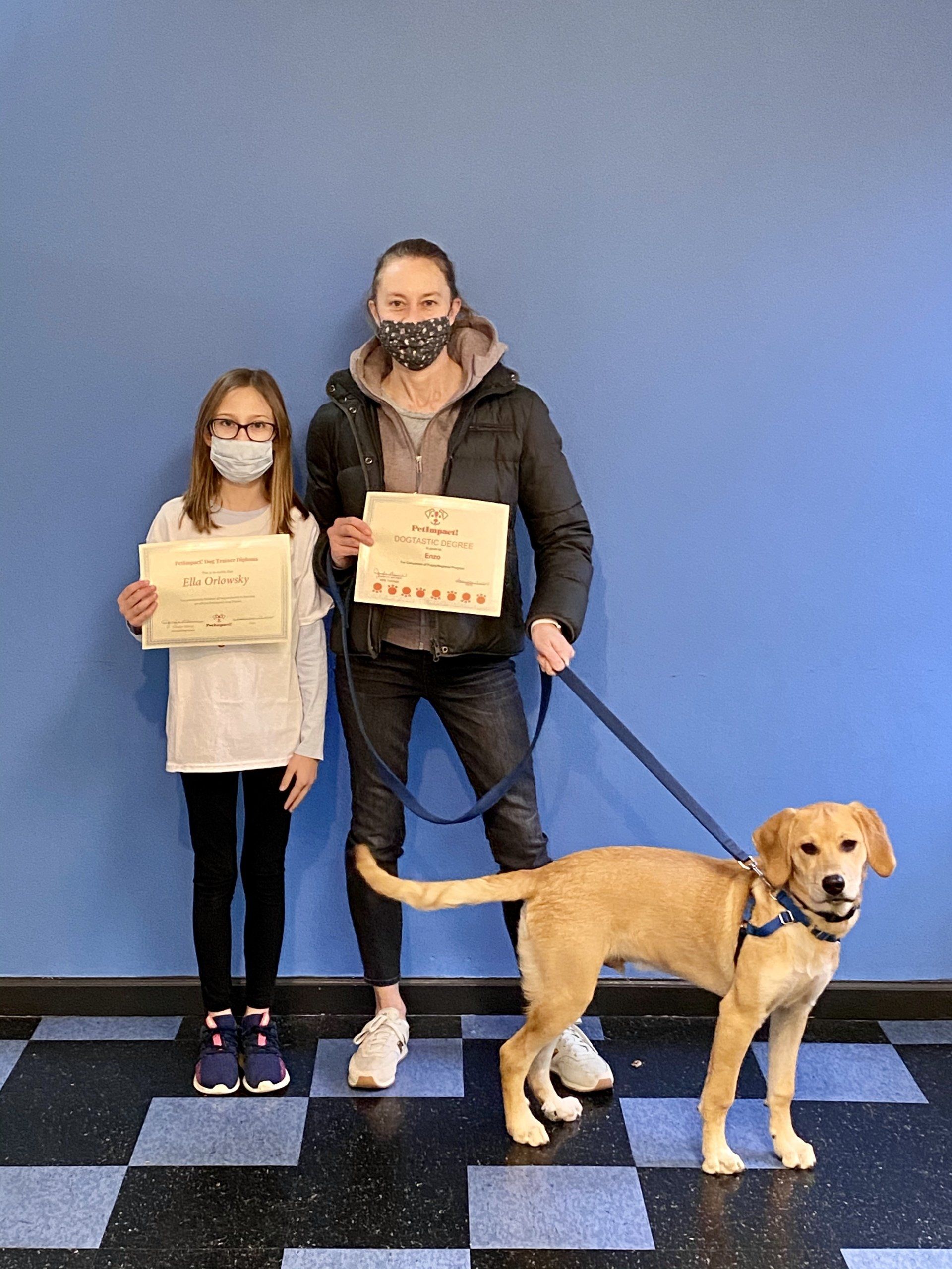 Woman, girl, and dog pose in front of a blue wall holding certificates.