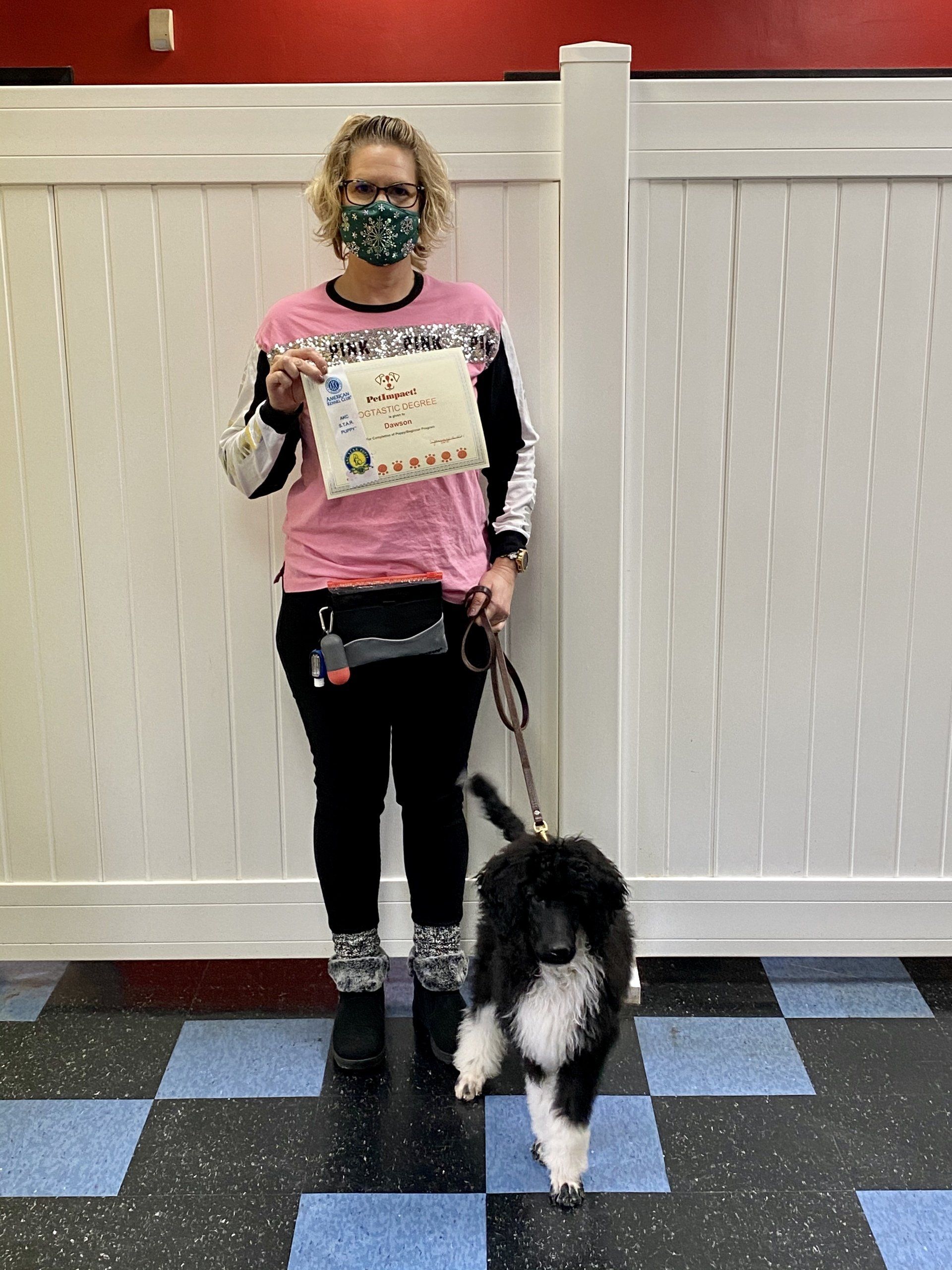 Woman and black and white poodle, holding certificate, standing next to white fence, checkered floor.