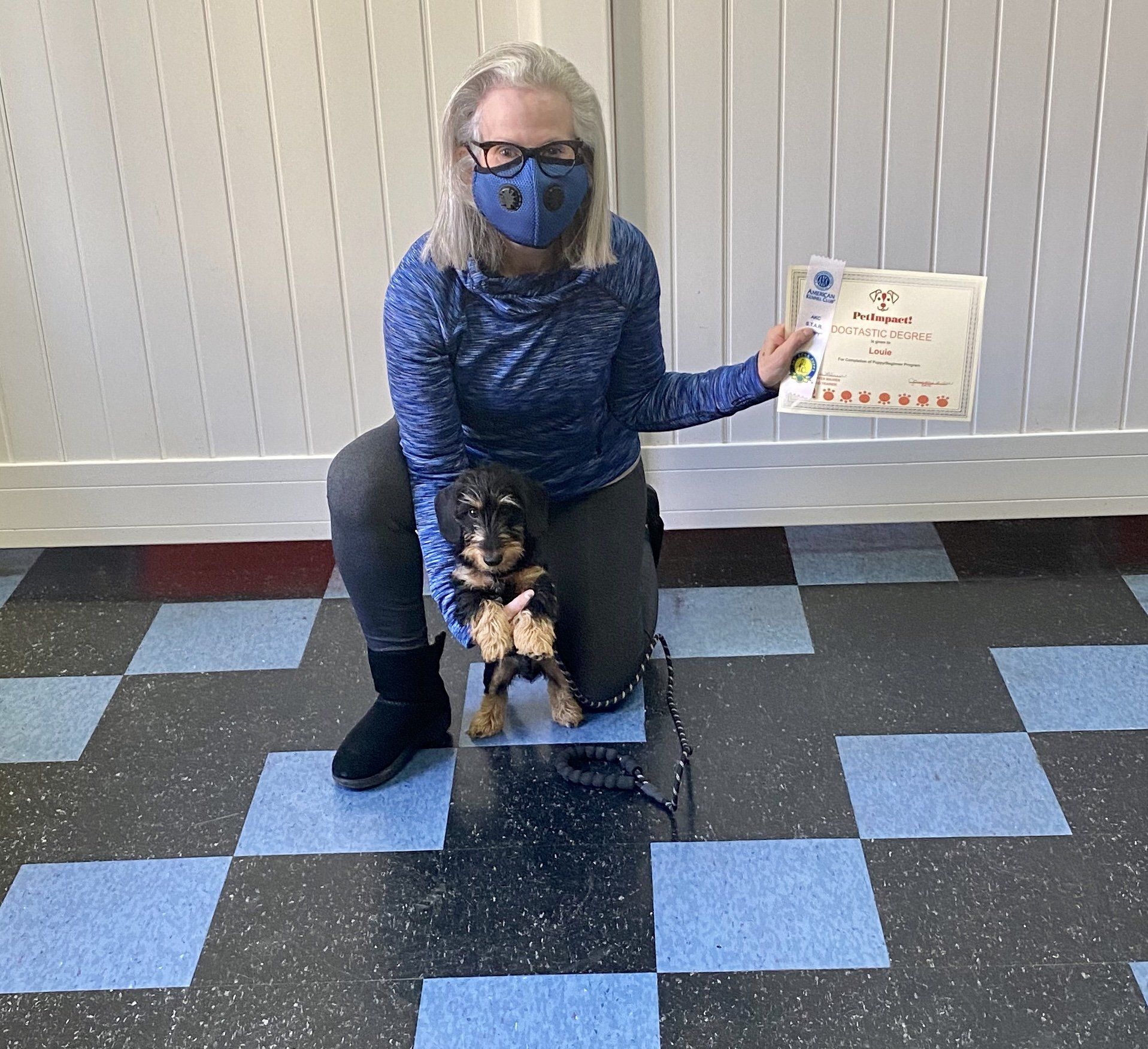 Woman in mask kneels with small dog and a certificate, on a blue and black tiled floor.