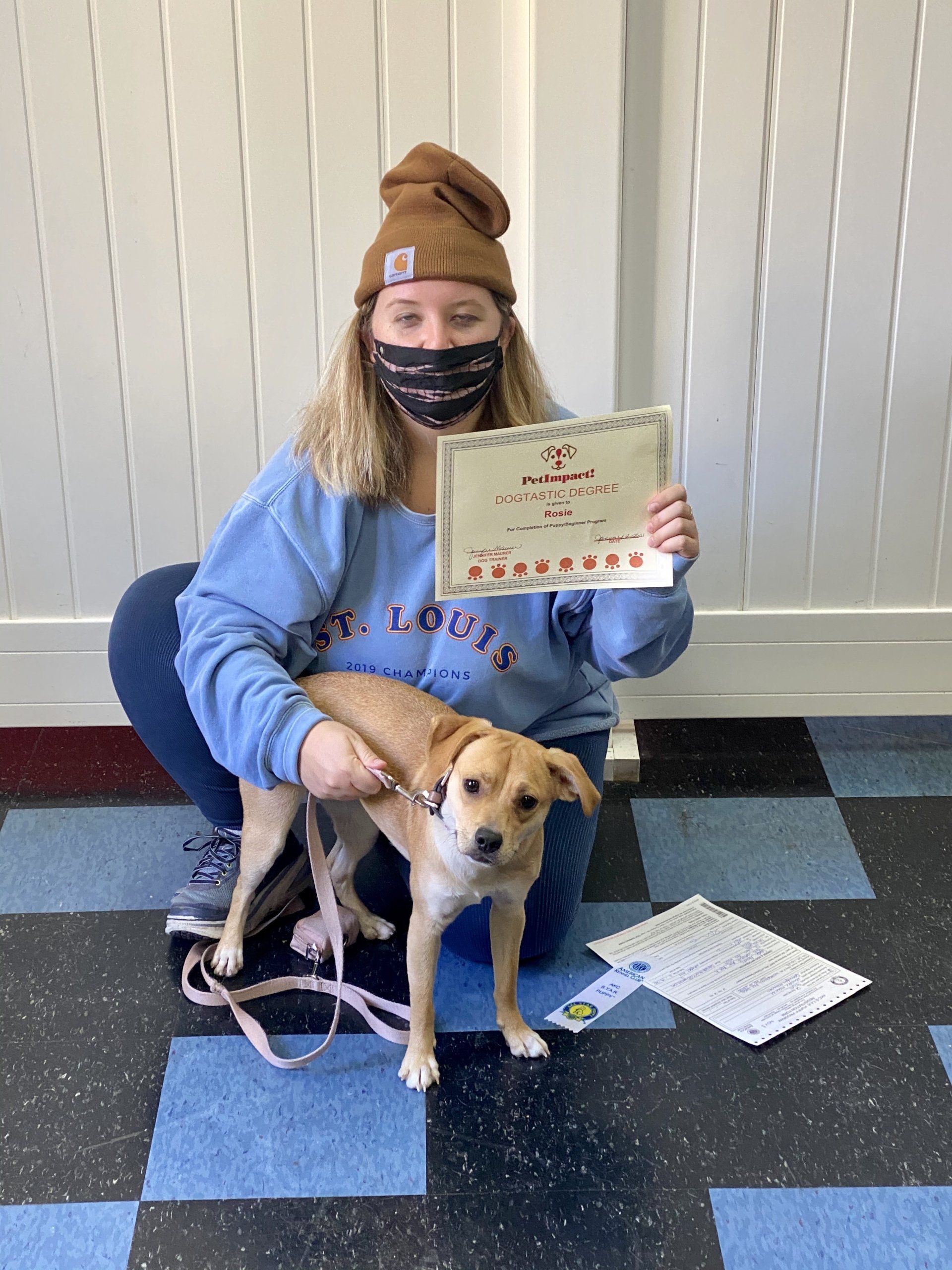 Woman in blue sweatshirt, beanie, and mask with dog, holding certificate. Sitting on blue checkered floor.