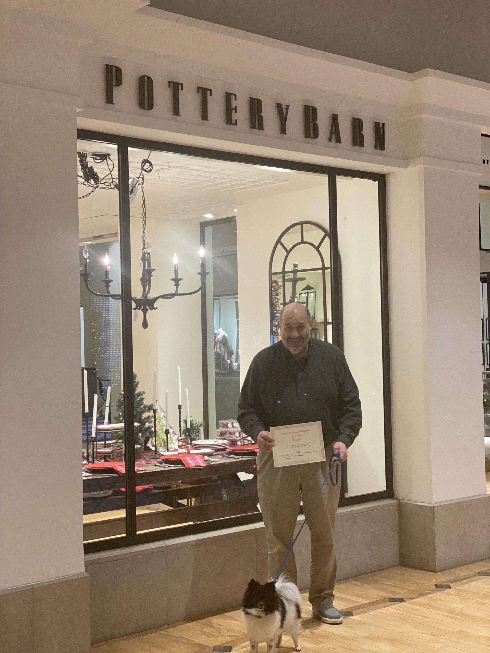 Man holding paper, with small dog, in front of Pottery Barn store.
