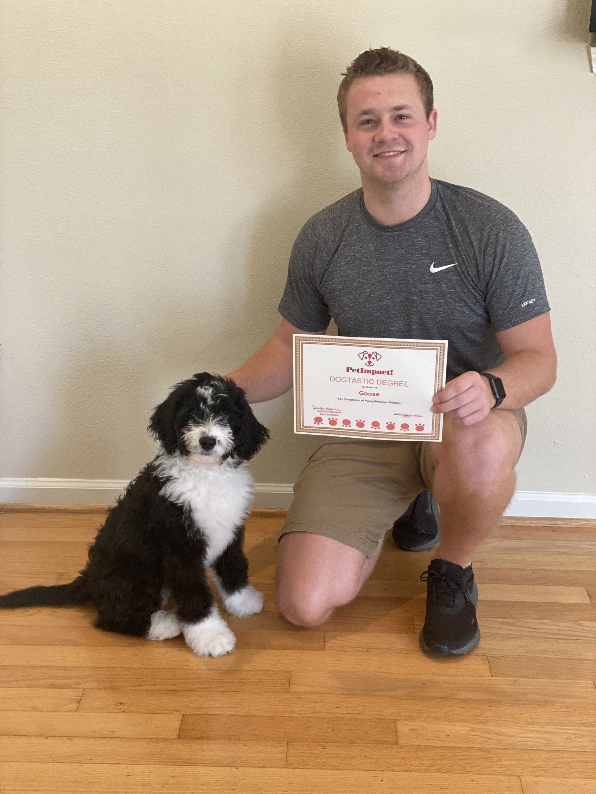 Man kneeling next to a black and white dog, holding a certificate. Wooden floor, neutral wall.