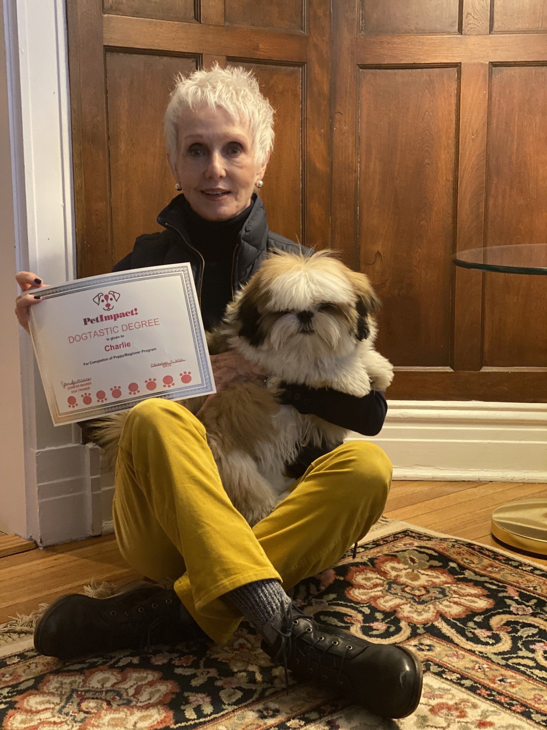 Woman holding a Shih Tzu and certificate, sitting on a rug near a wooden door; she wears yellow pants, a black jacket, and boots.