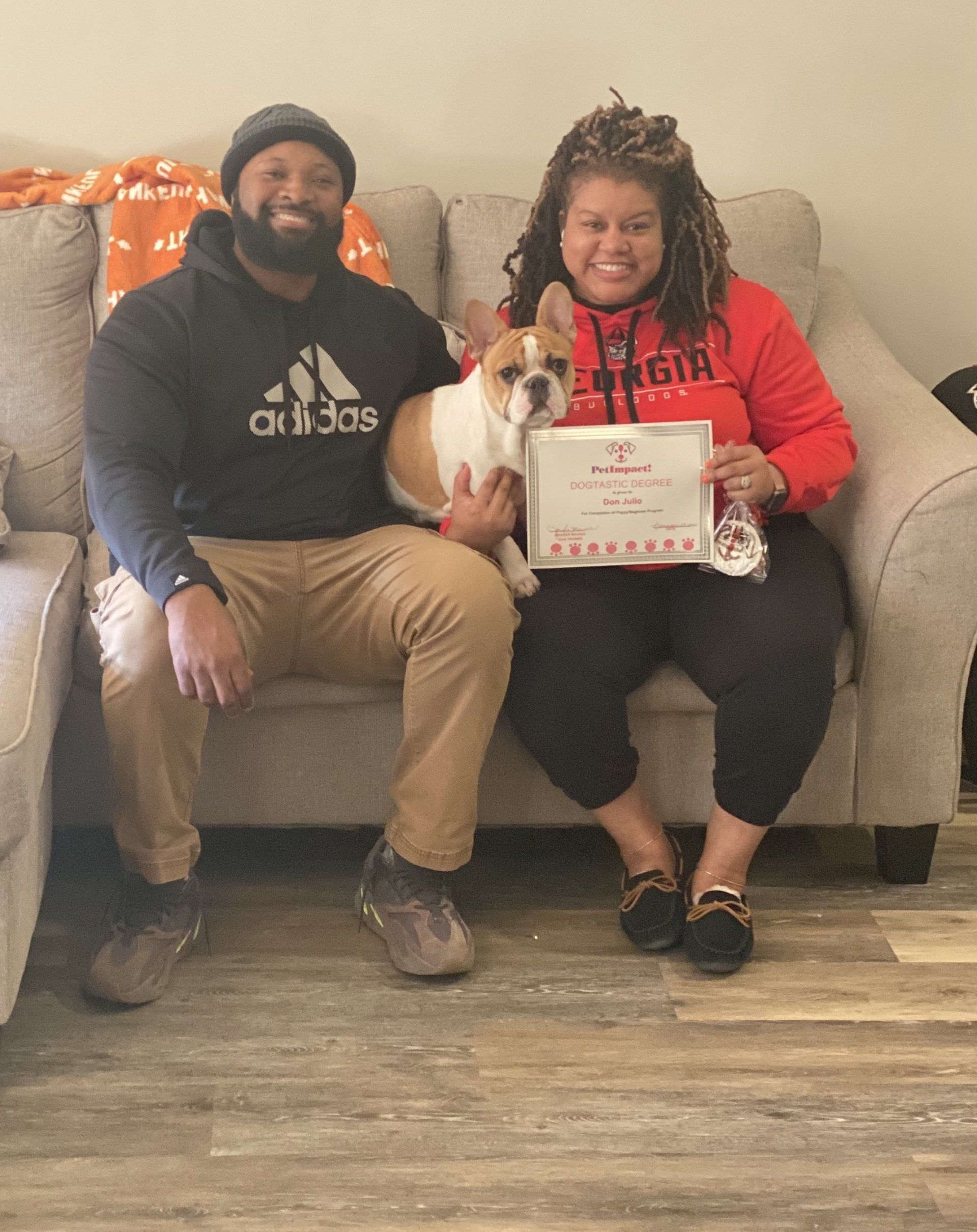 Couple on couch with dog, holding certificate. The man wears an Adidas hoodie, the woman a red shirt.