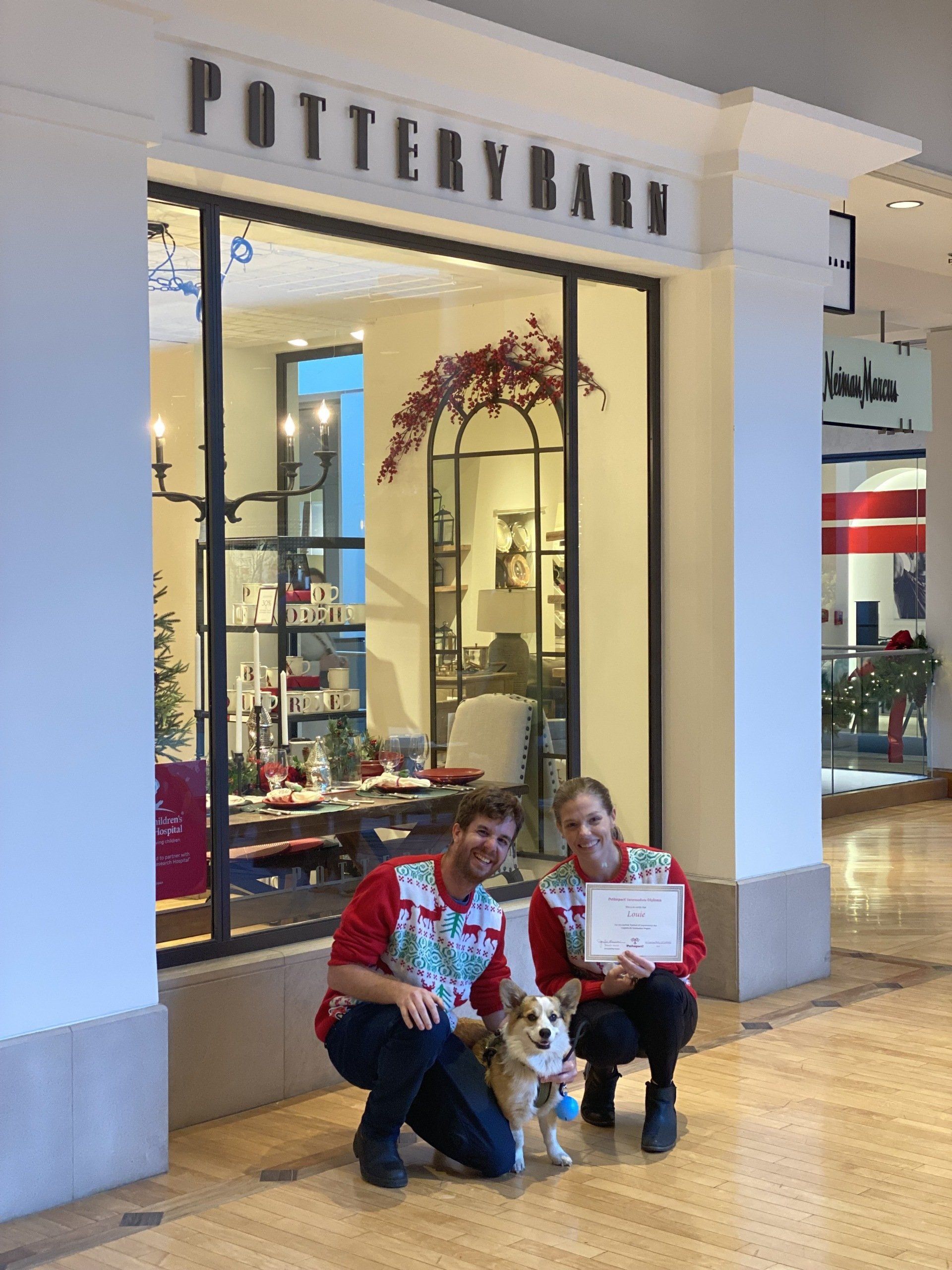 Two people and a dog in front of Pottery Barn store. They hold a paper. Store window with holiday decor.