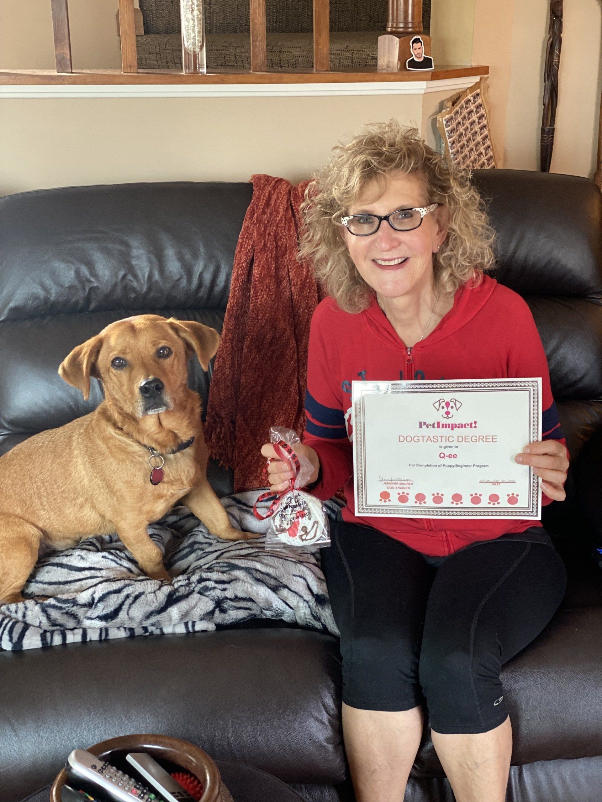 Woman and dog on a couch holding a framed award. Woman smiling in red and black, dog looks at the camera.