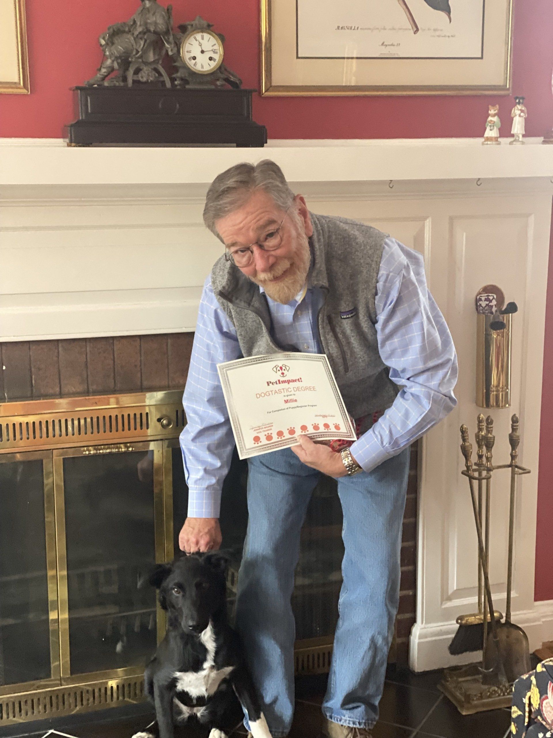 Man in glasses, holding framed document, petting black and white dog by fireplace.