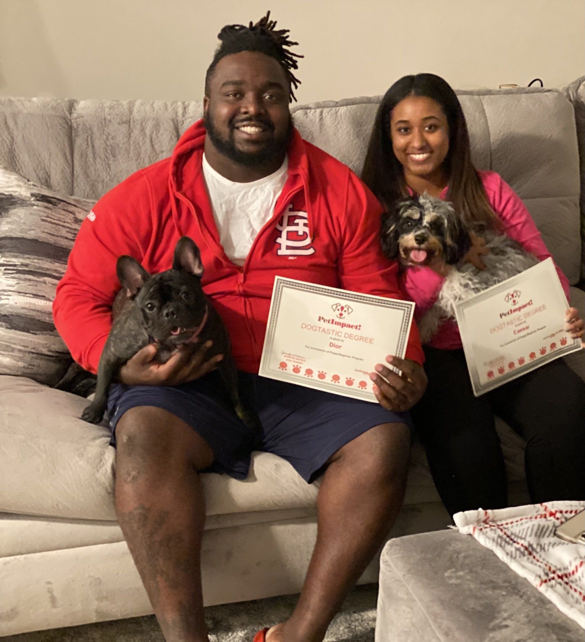 Couple holding dogs and certificates on a couch; man in red zip-up jacket, woman in pink; dogs are black Frenchie and small, fluffy, gray.