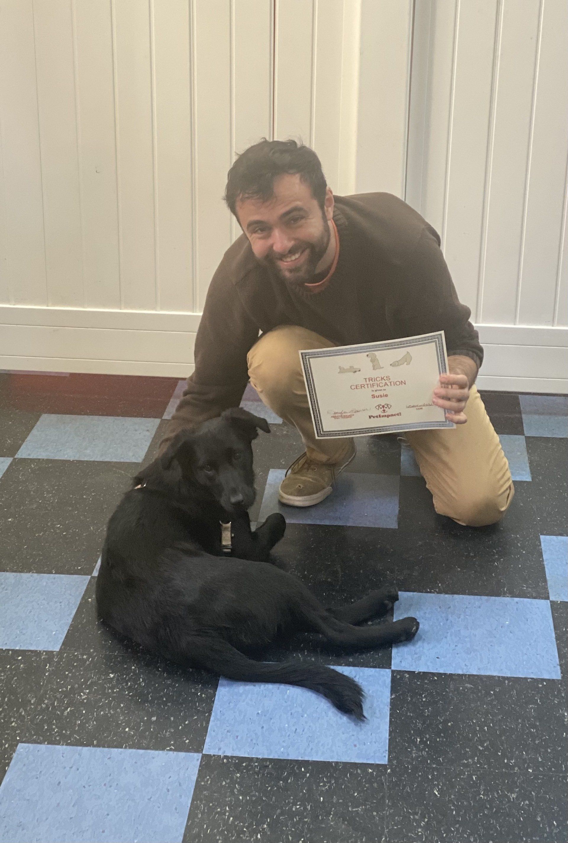 Man kneeling with dog, holding a certificate; they're on a checkered floor with a light wall backdrop.