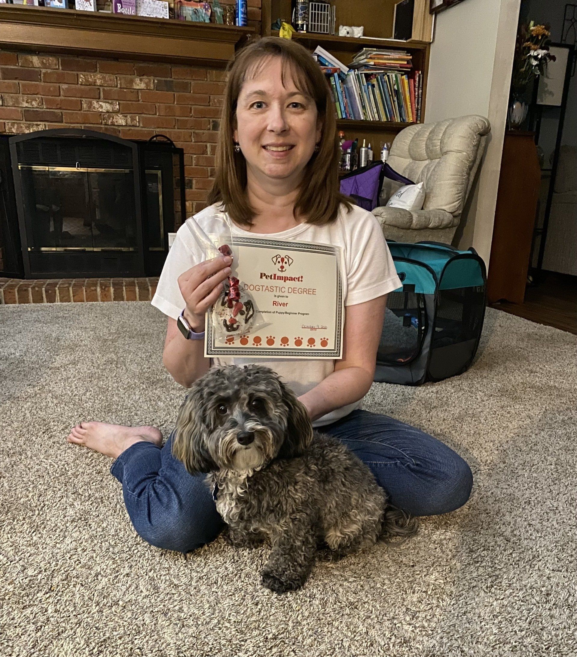 Woman sits with a dog, holding a certificate by a fireplace.