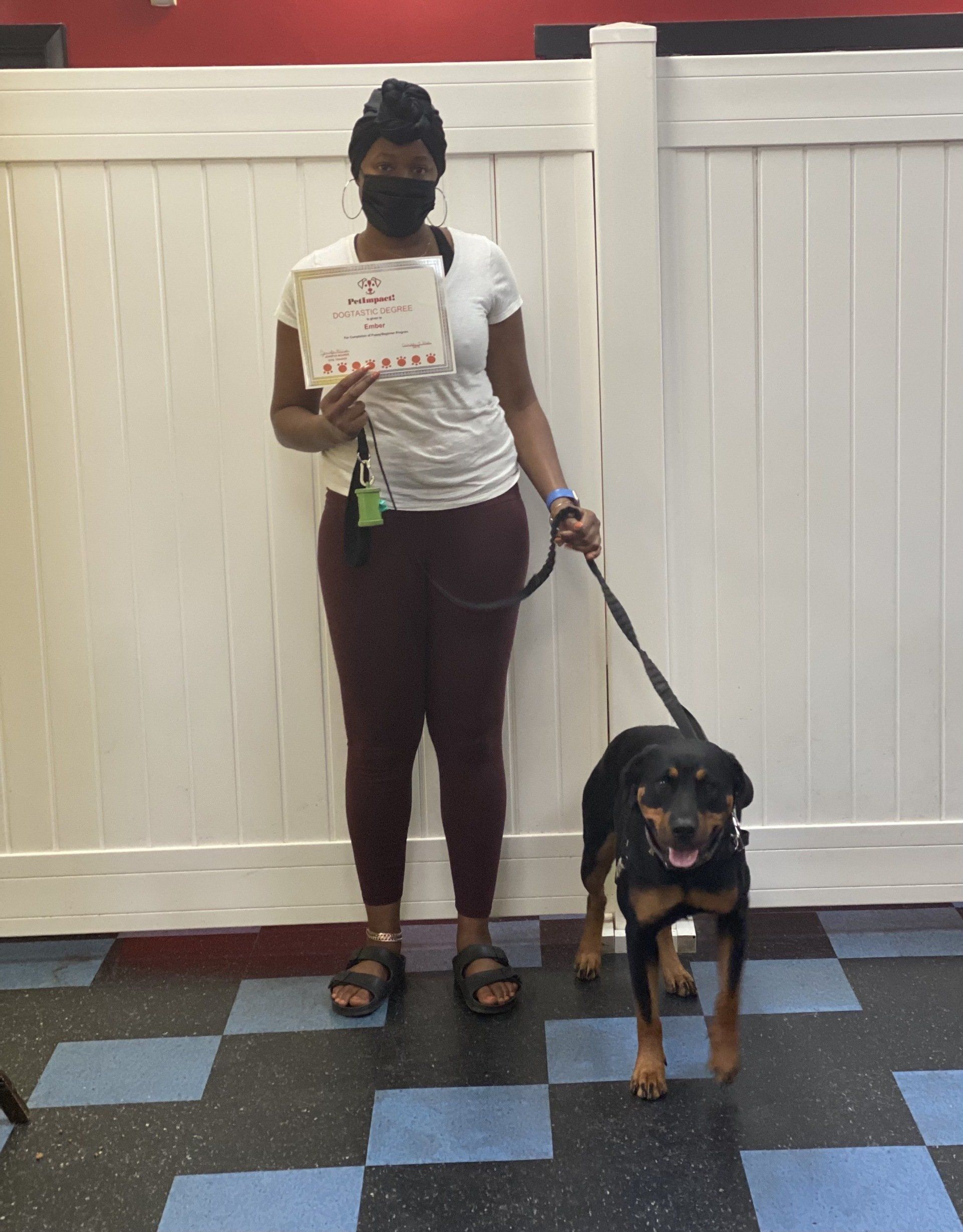 Woman in mask holds certificate, stands with a dog on a leash in front of a white fence.