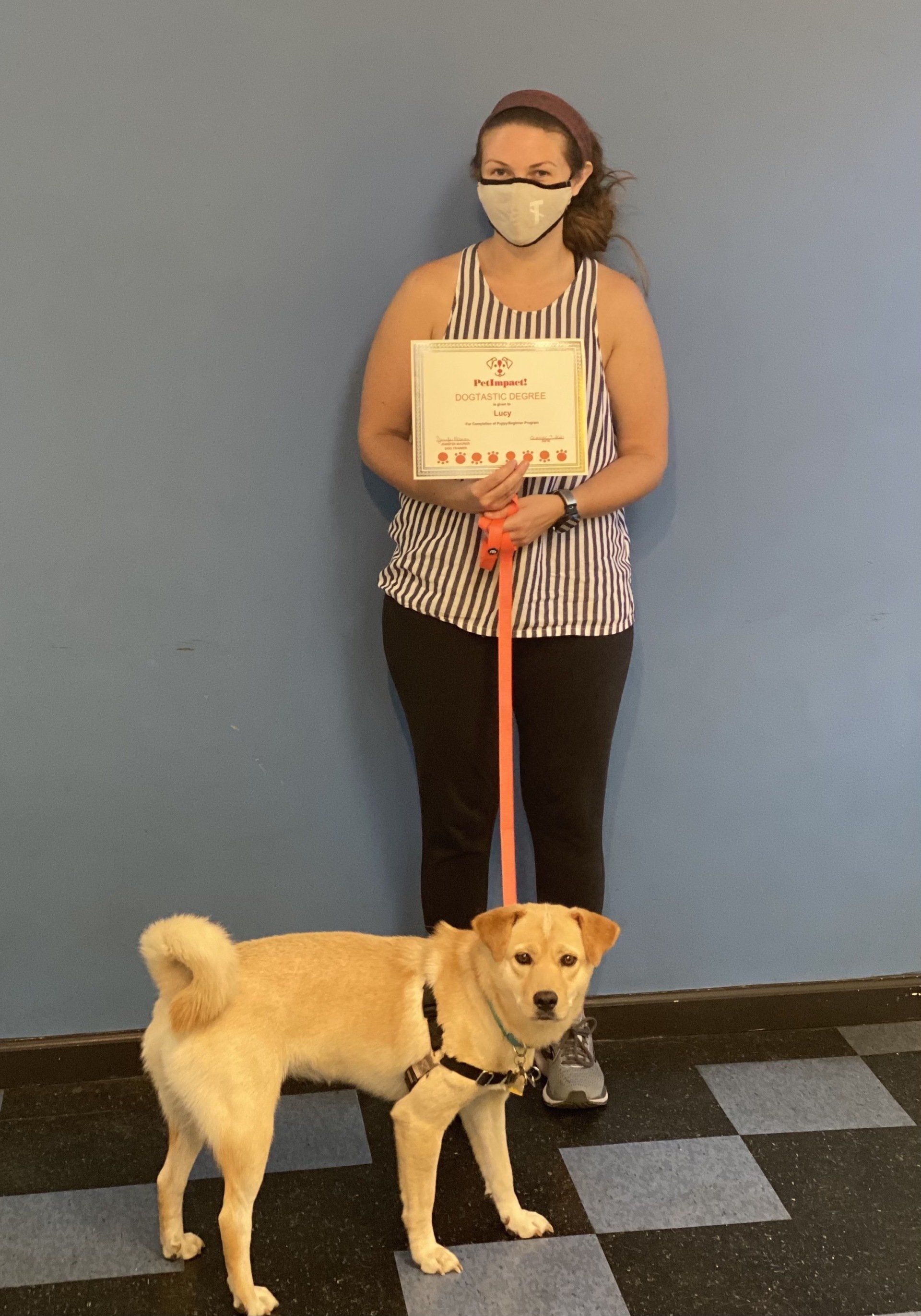 Woman in mask holds certificate, stands with a light-brown dog on leash, in front of a blue wall.