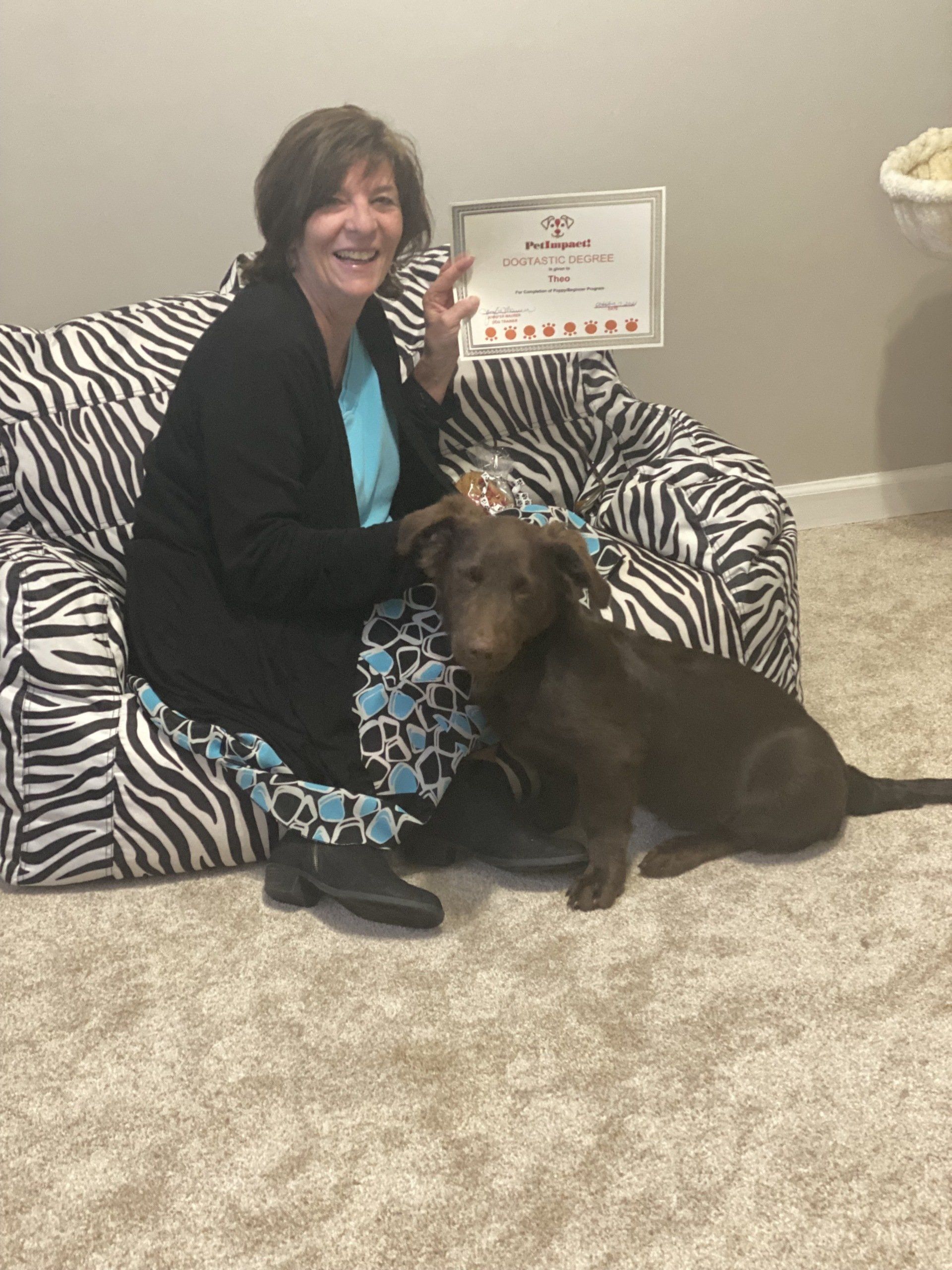 Woman and brown dog seated on zebra-print beanbag, holding a framed document and smiling.