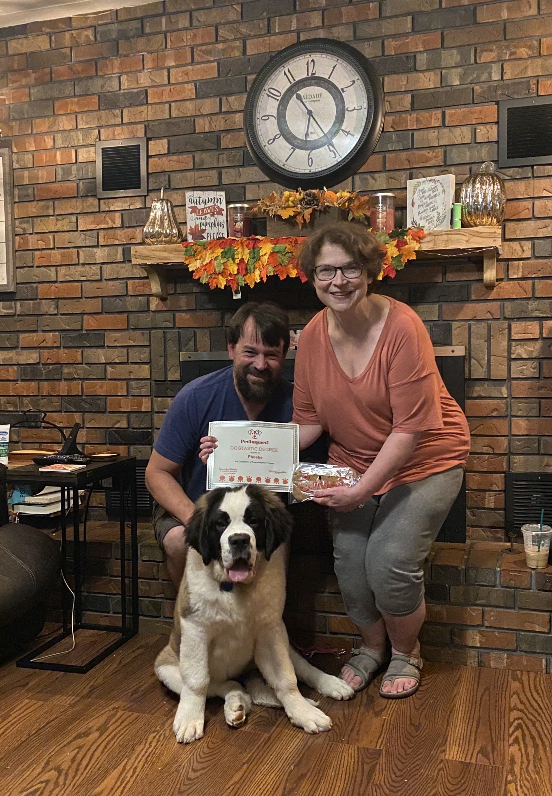 A couple and a St. Bernard dog hold a certificate and cake in front of a brick fireplace, decorated with fall foliage.