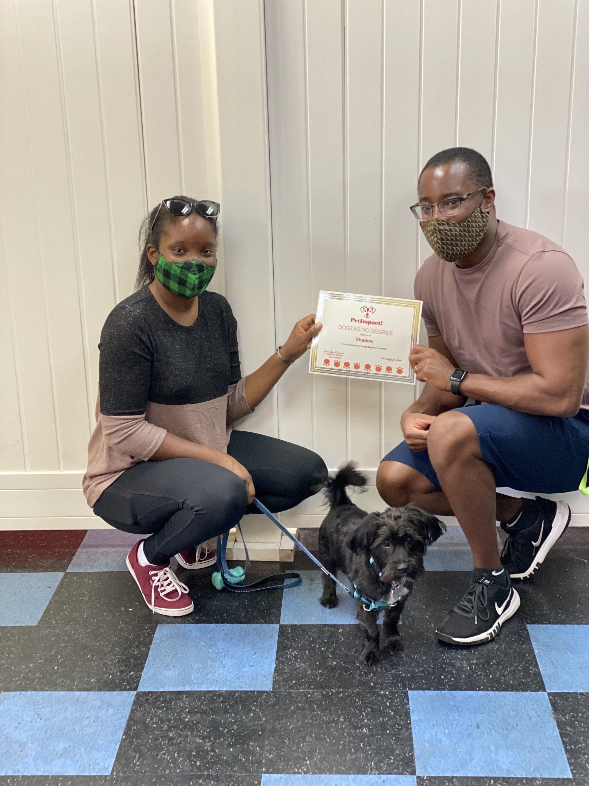Two people in masks pose with a small dog and a box indoors.