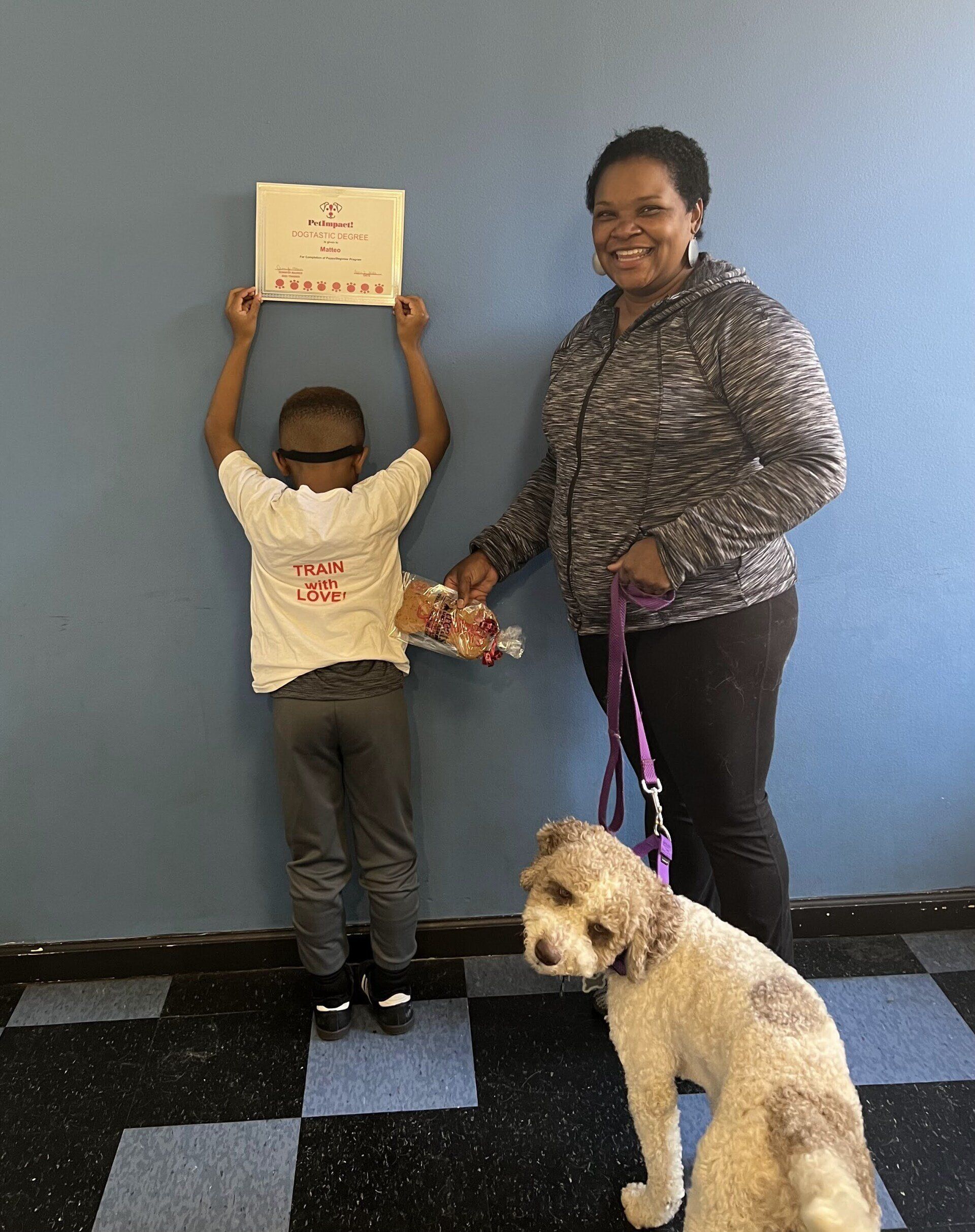 A child holds a certificate; a woman with a dog looks on. The dog is on a leash. All are indoors.