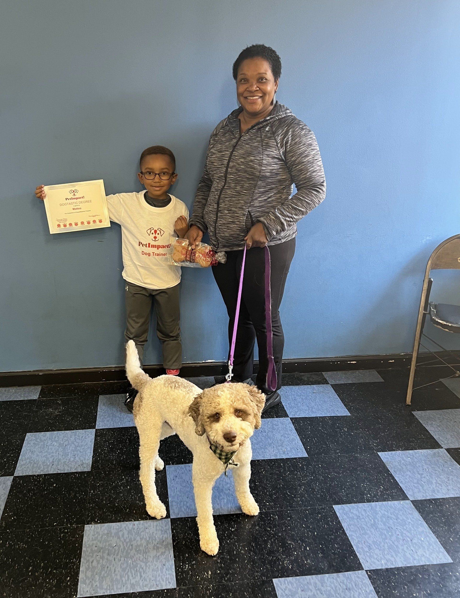 A woman and child stand with a dog. The child holds a sign and treats. All are in a room with a blue wall.