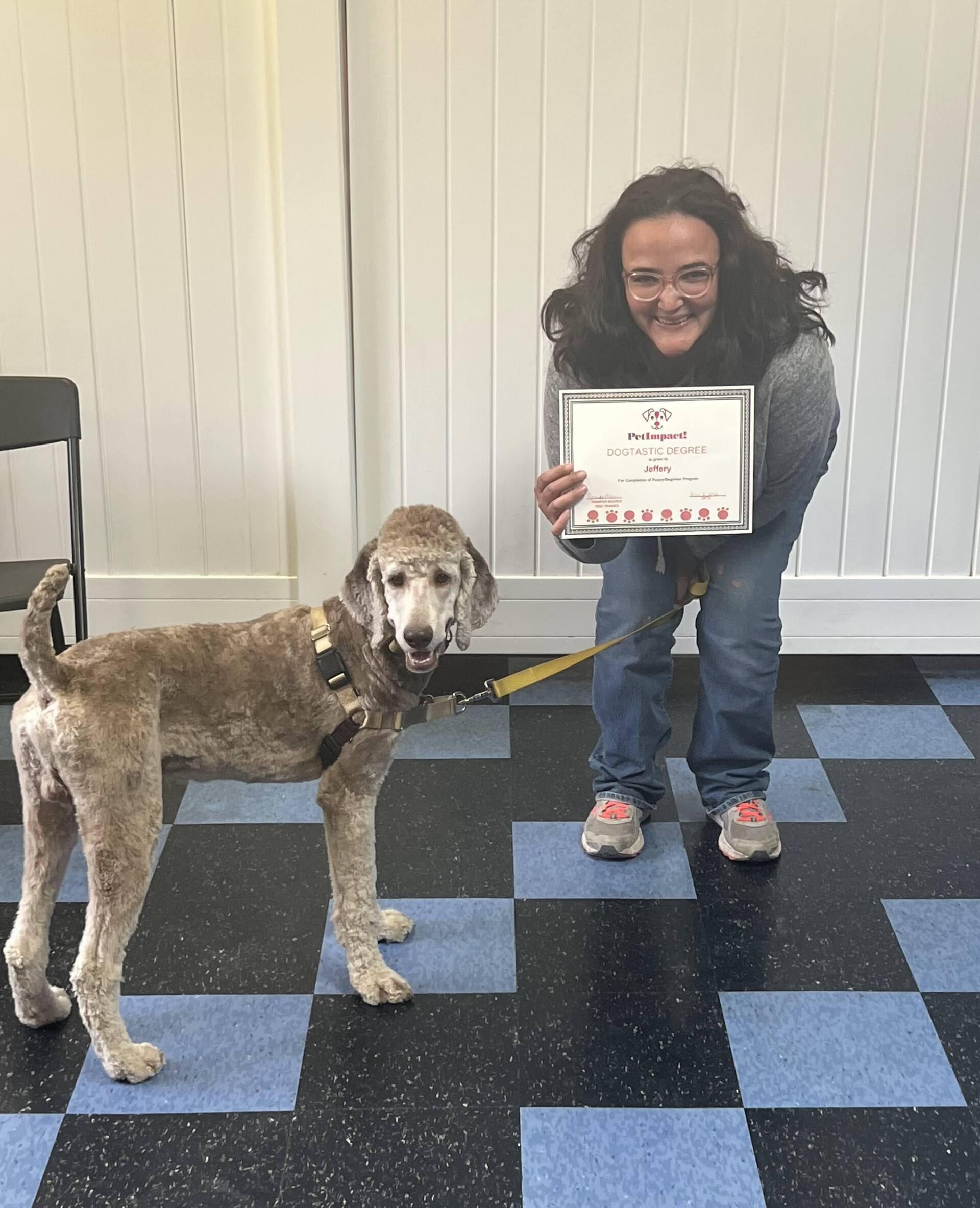 Dog and person posing, dog on leash. Person holds certificate, smiles. Blue and black checkered floor.