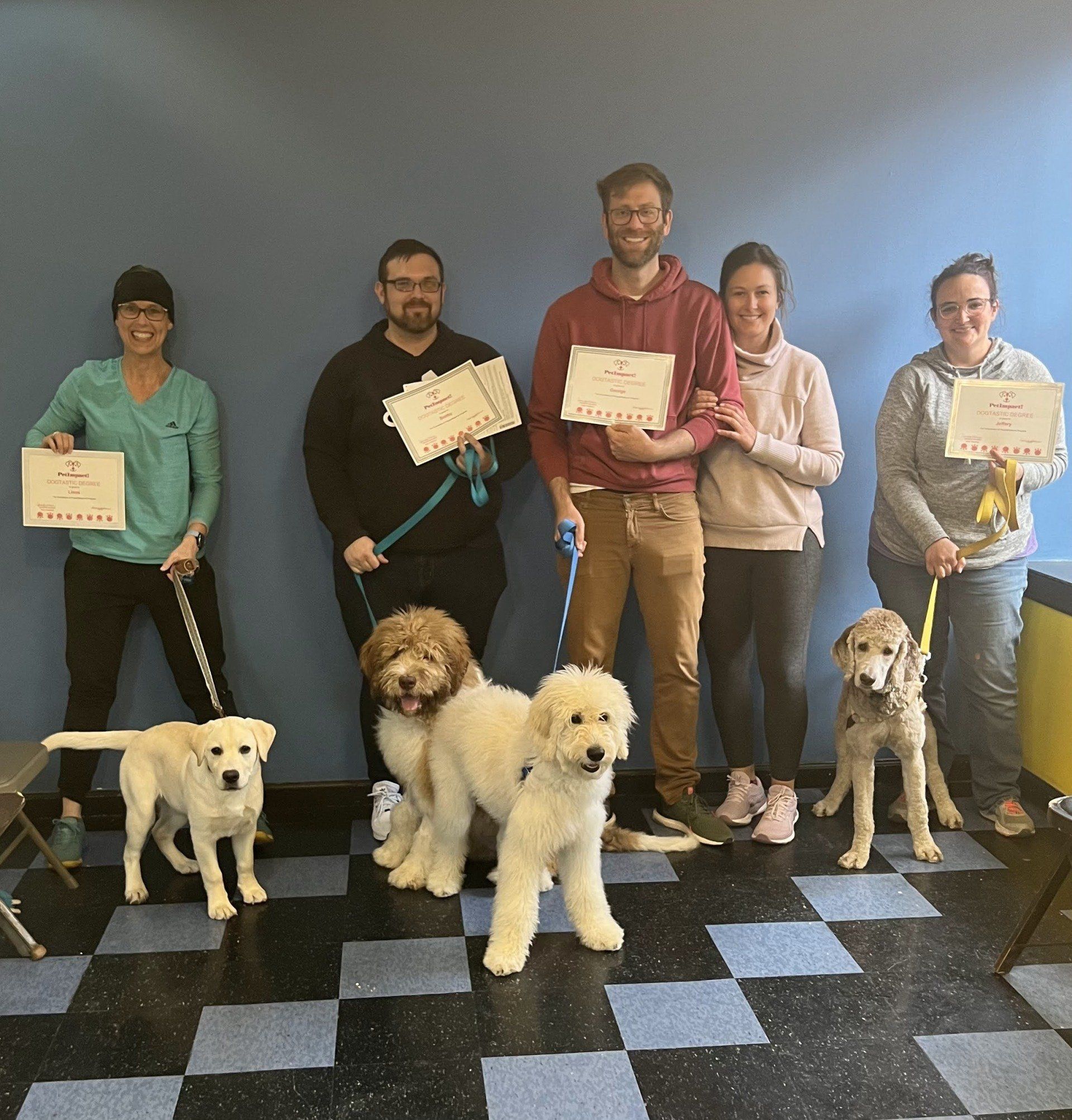 People with dogs hold certificates, posing in front of a blue wall and checkered floor.