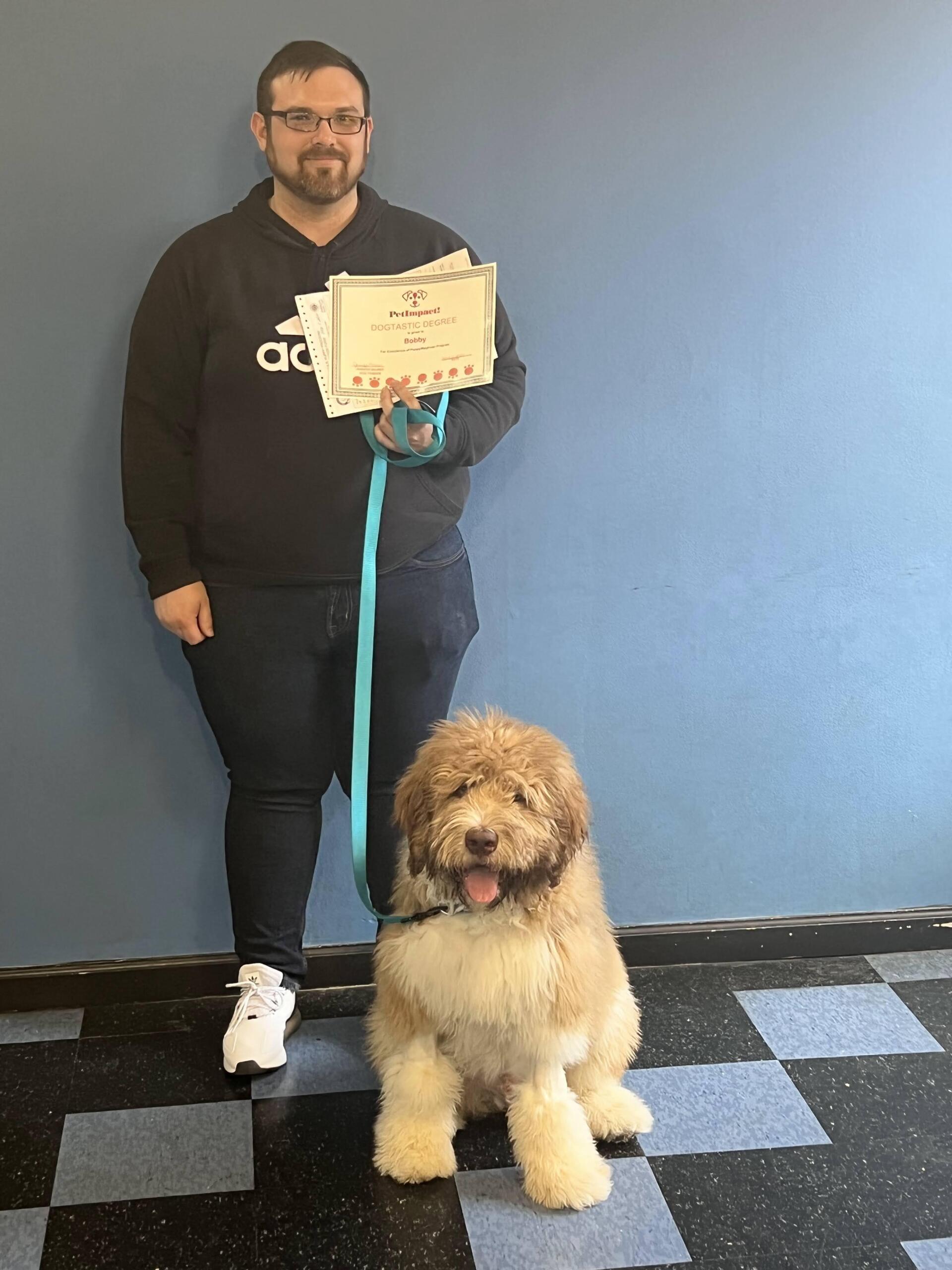Man holding papers with a light brown dog on a leash; dog is sitting. They are in front of a blue wall and black and white floor.