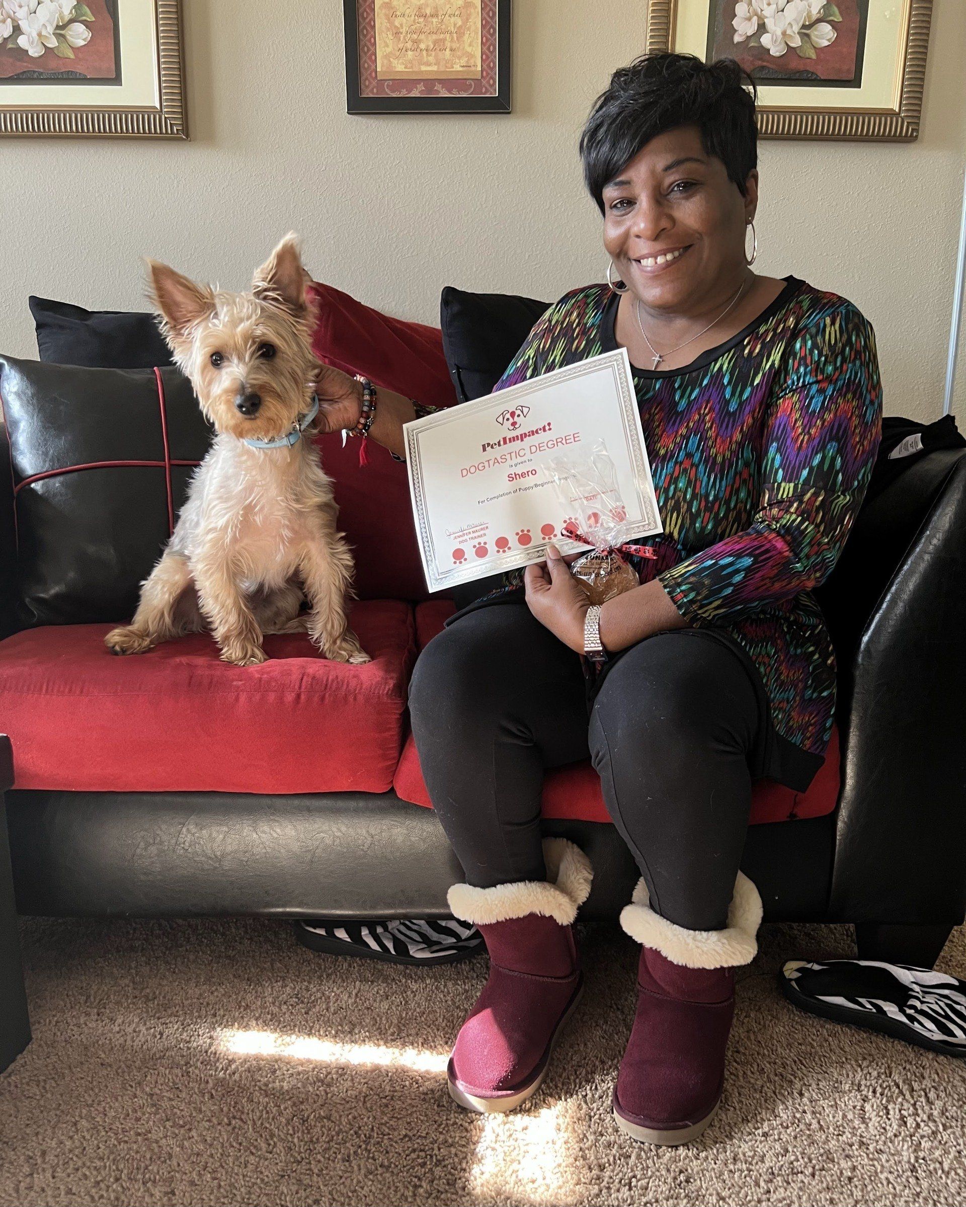 Woman holding certificate, smiling, seated on red couch with dog. Dog sits beside her. Indoors.