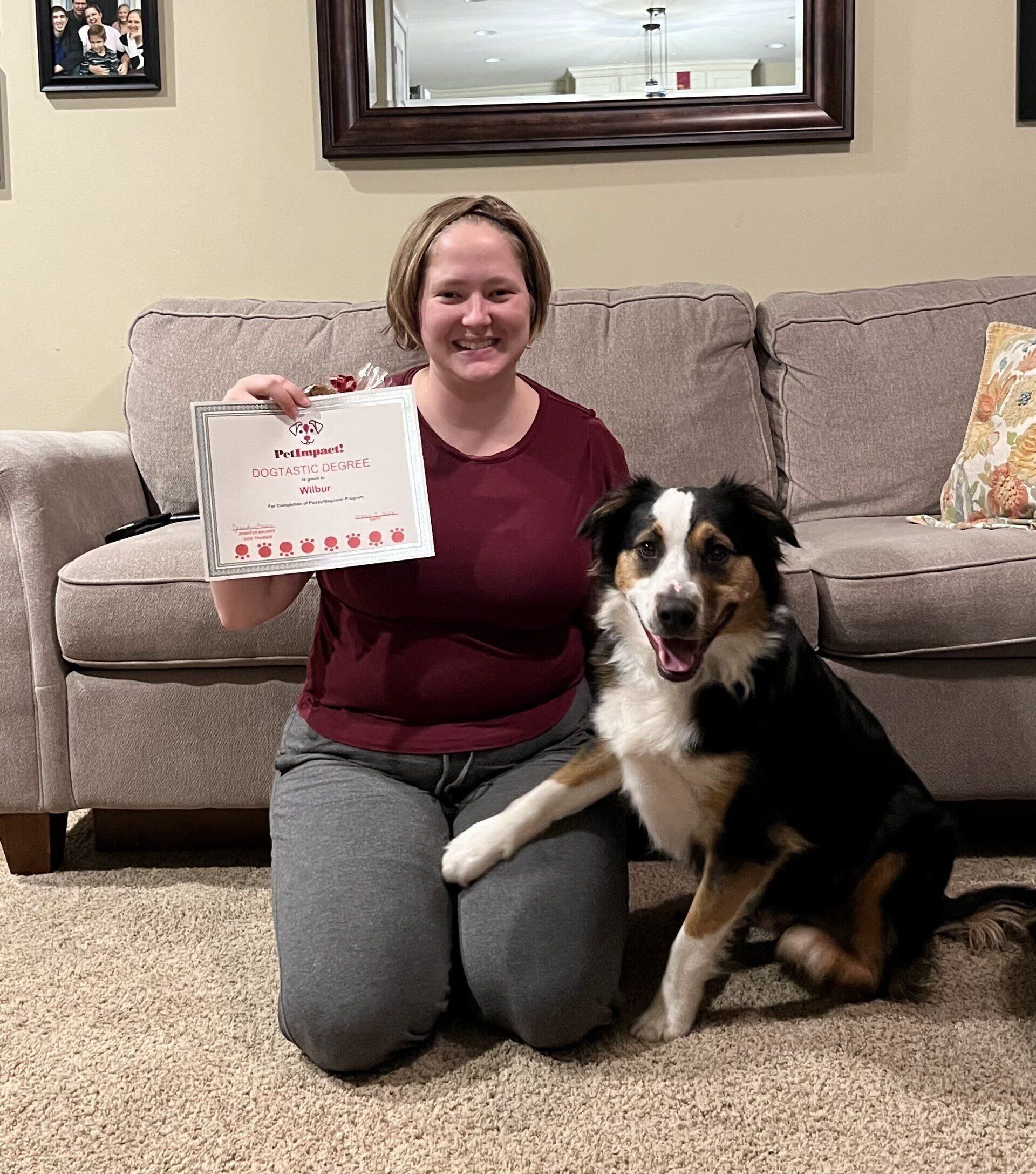 Woman kneeling with dog, holding certificate. Red shirt, gray pants. Dog is black, white, and tan. Living room setting.
