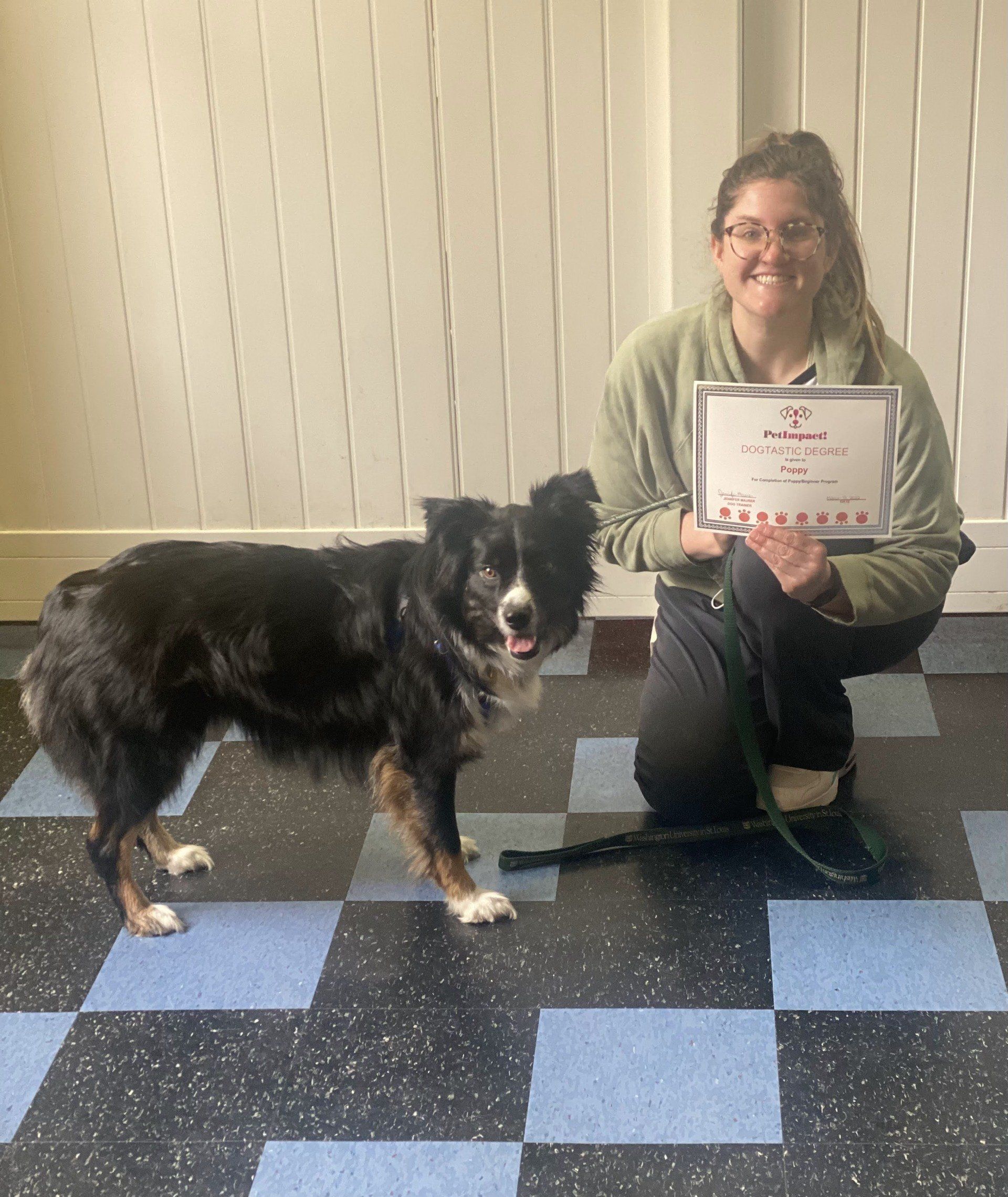 Woman kneeling next to dog, holding certificate. Dog is black and white. Tile floor. White wall.