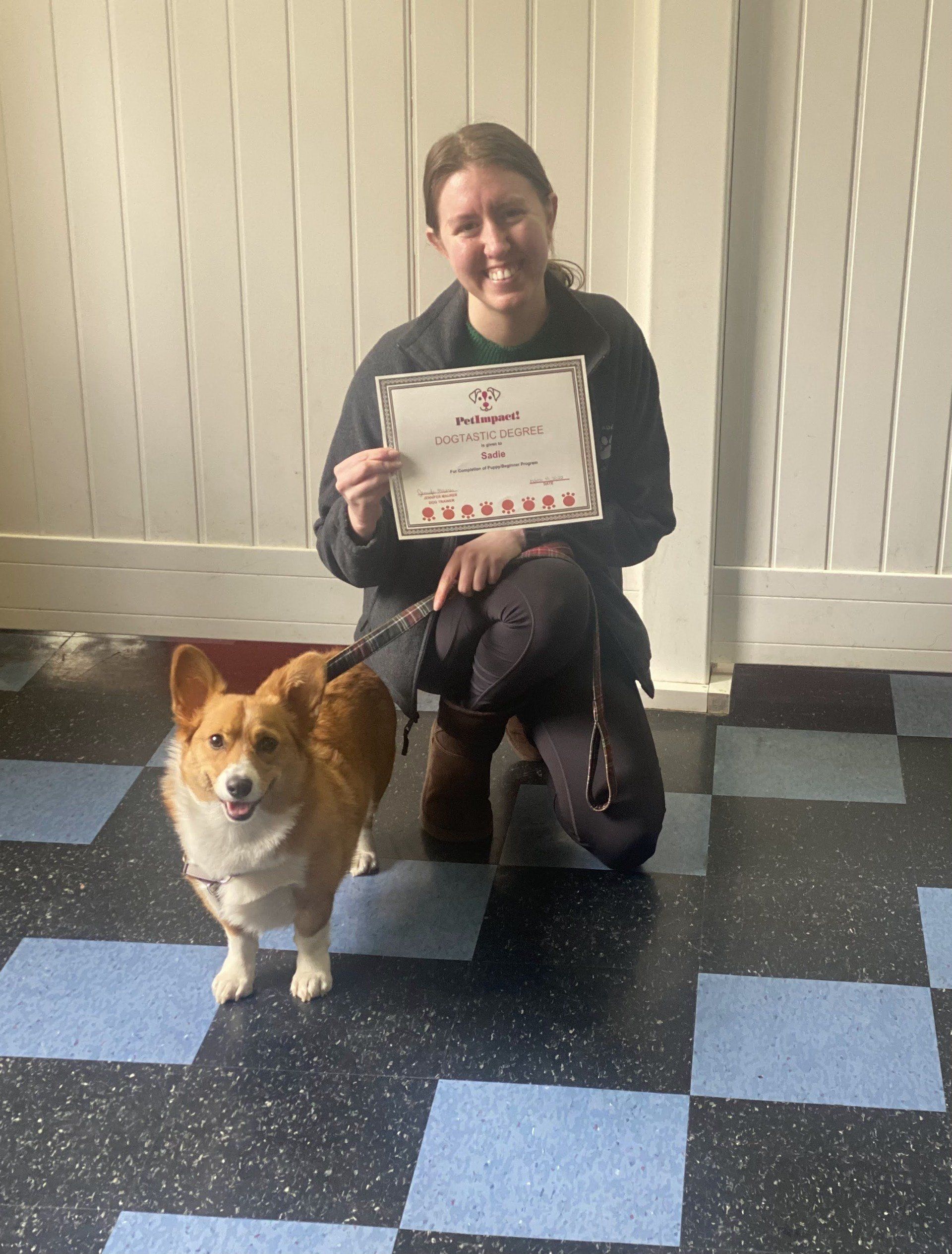 Woman and corgi dog pose with a certificate. The woman smiles, kneeling. Corgi wears a leash, standing on a patterned floor.