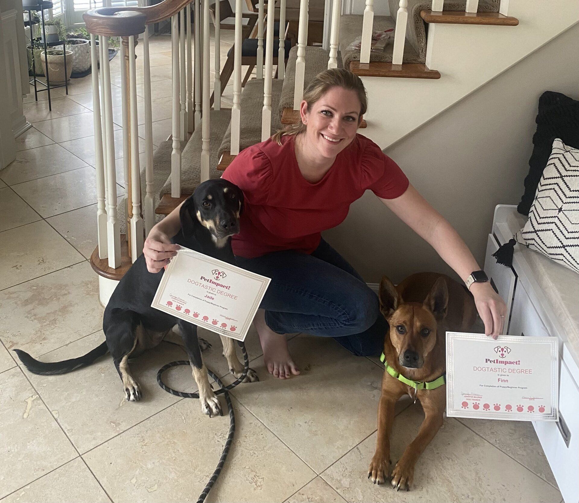 Woman kneels with two dogs, each holding a certificate. Indoors, smiles, stairwell visible.
