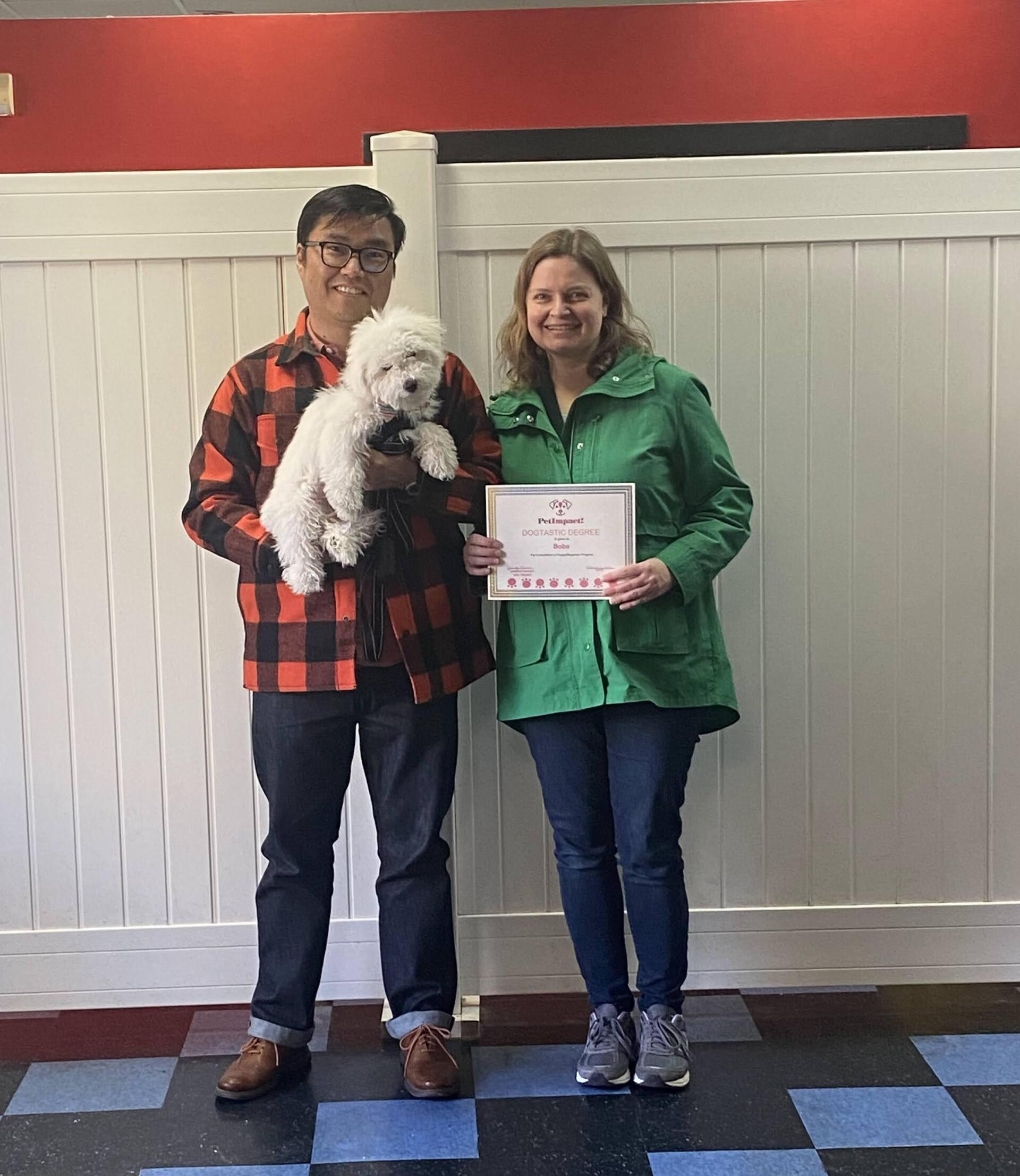 Two people hold a small white dog and a certificate in front of a white fence.