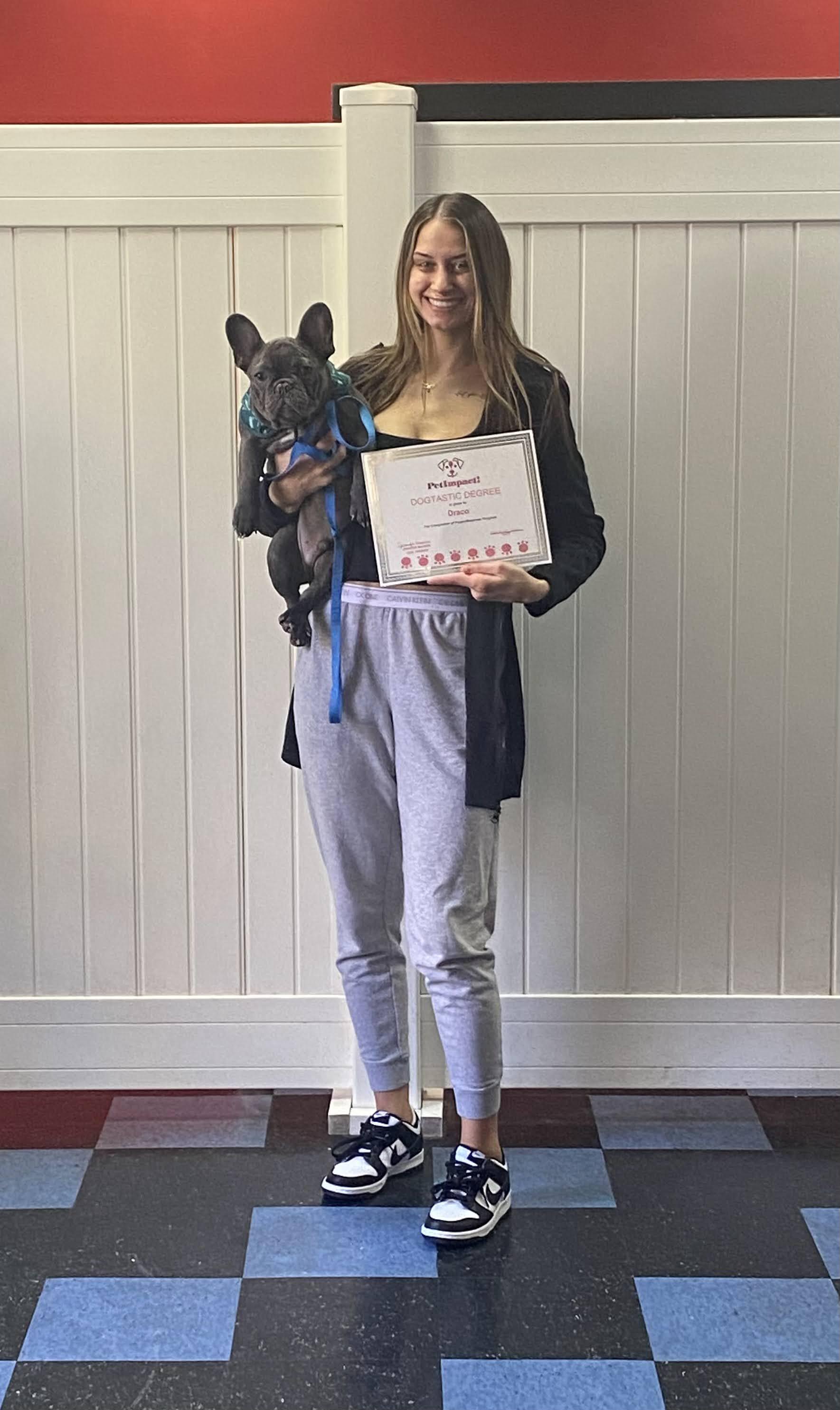 Woman holding a dog and a certificate stands against a white wall with a red stripe.