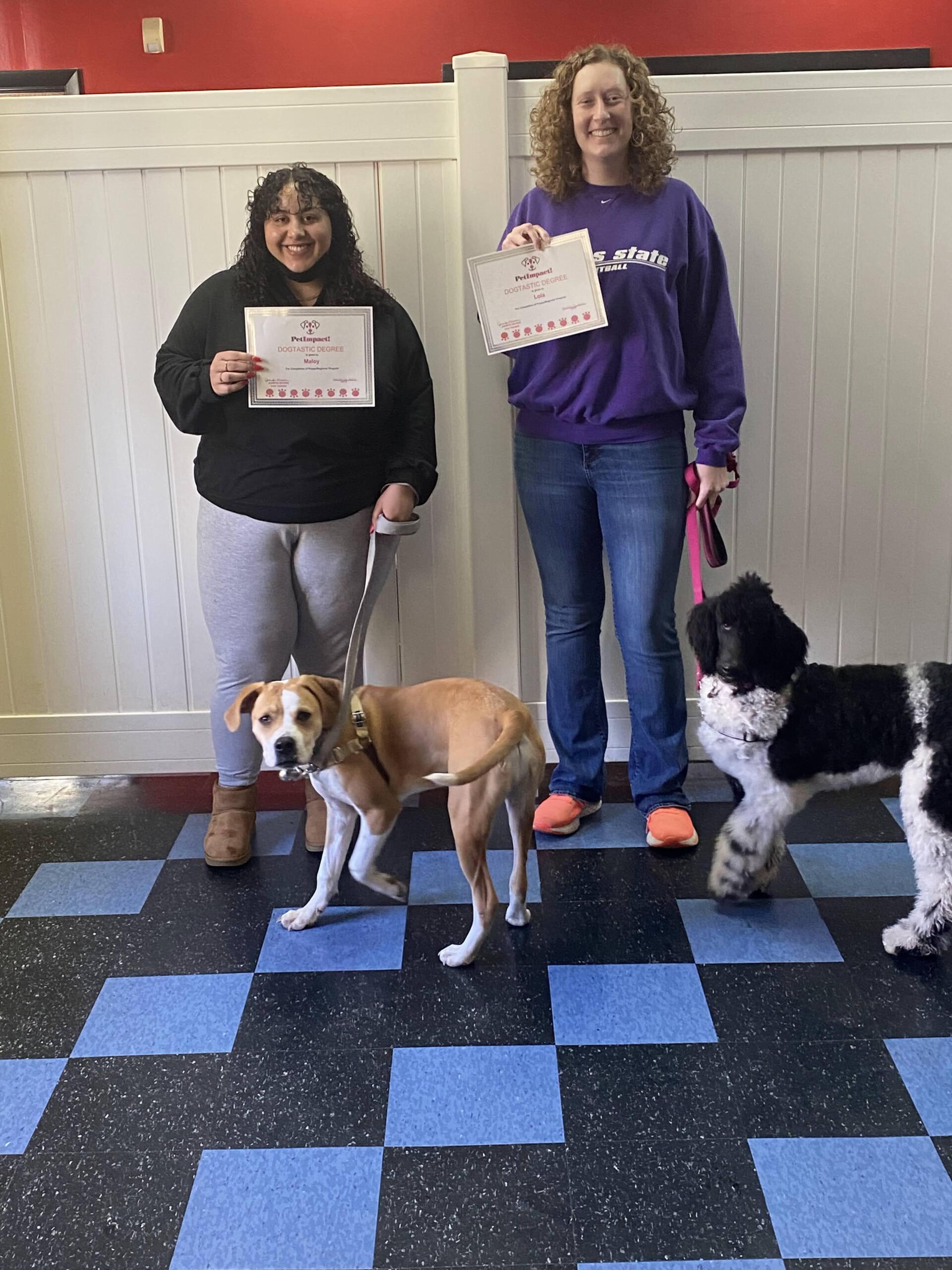 Two women with dogs, holding certificates, posing on a blue-tiled floor.