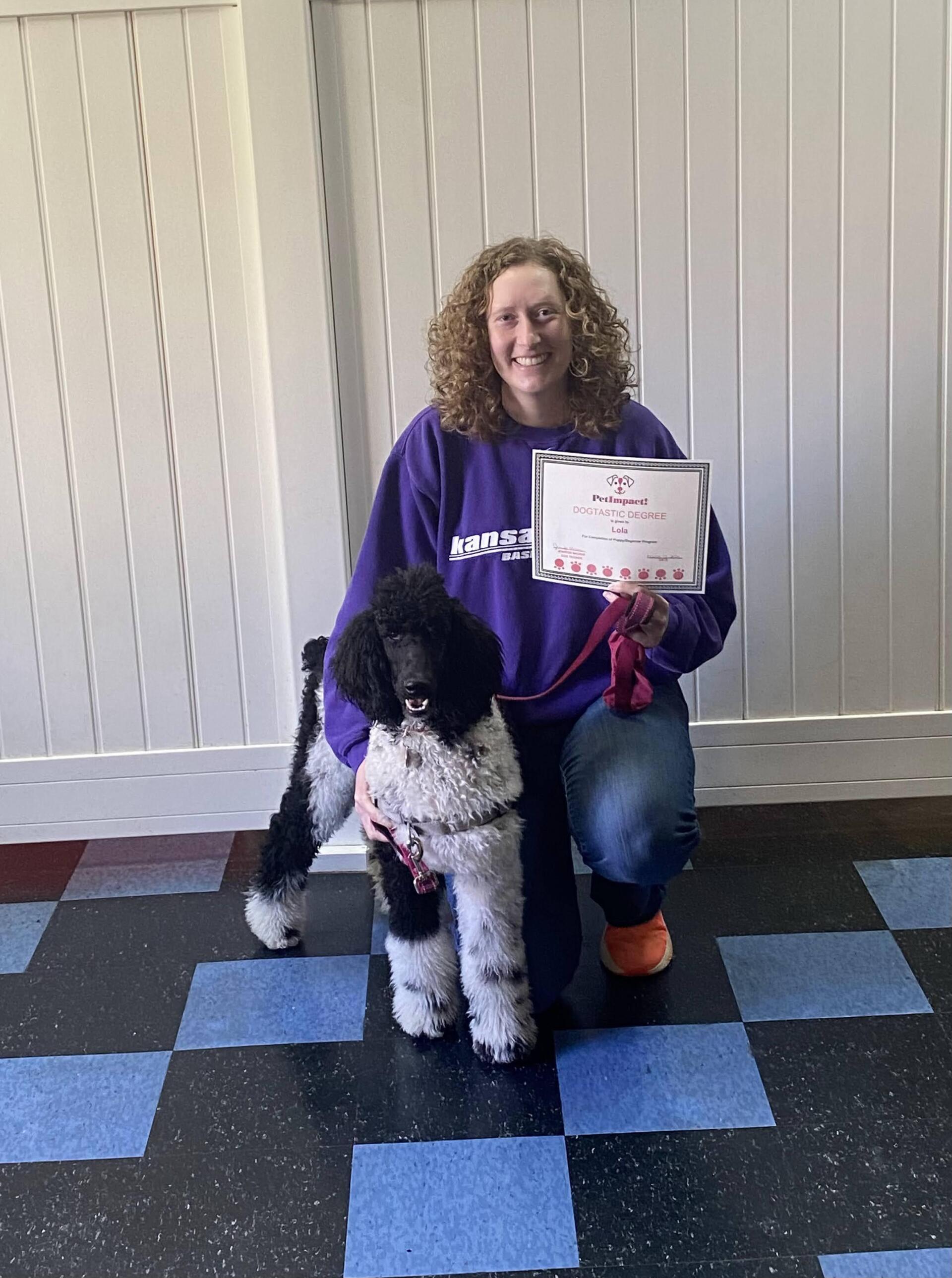 Woman kneeling with a black and white poodle, holding a certificate. Blue, black, and white checkered floor.