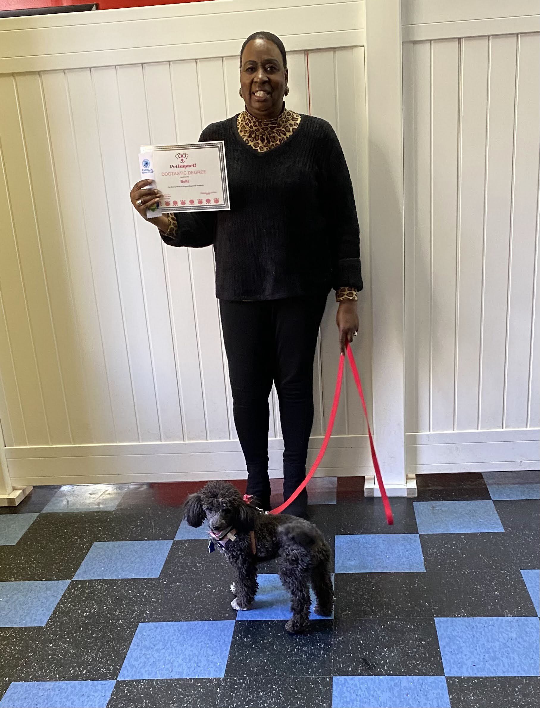 Woman holding certificate, standing with small black dog on a leash in front of a white fence.