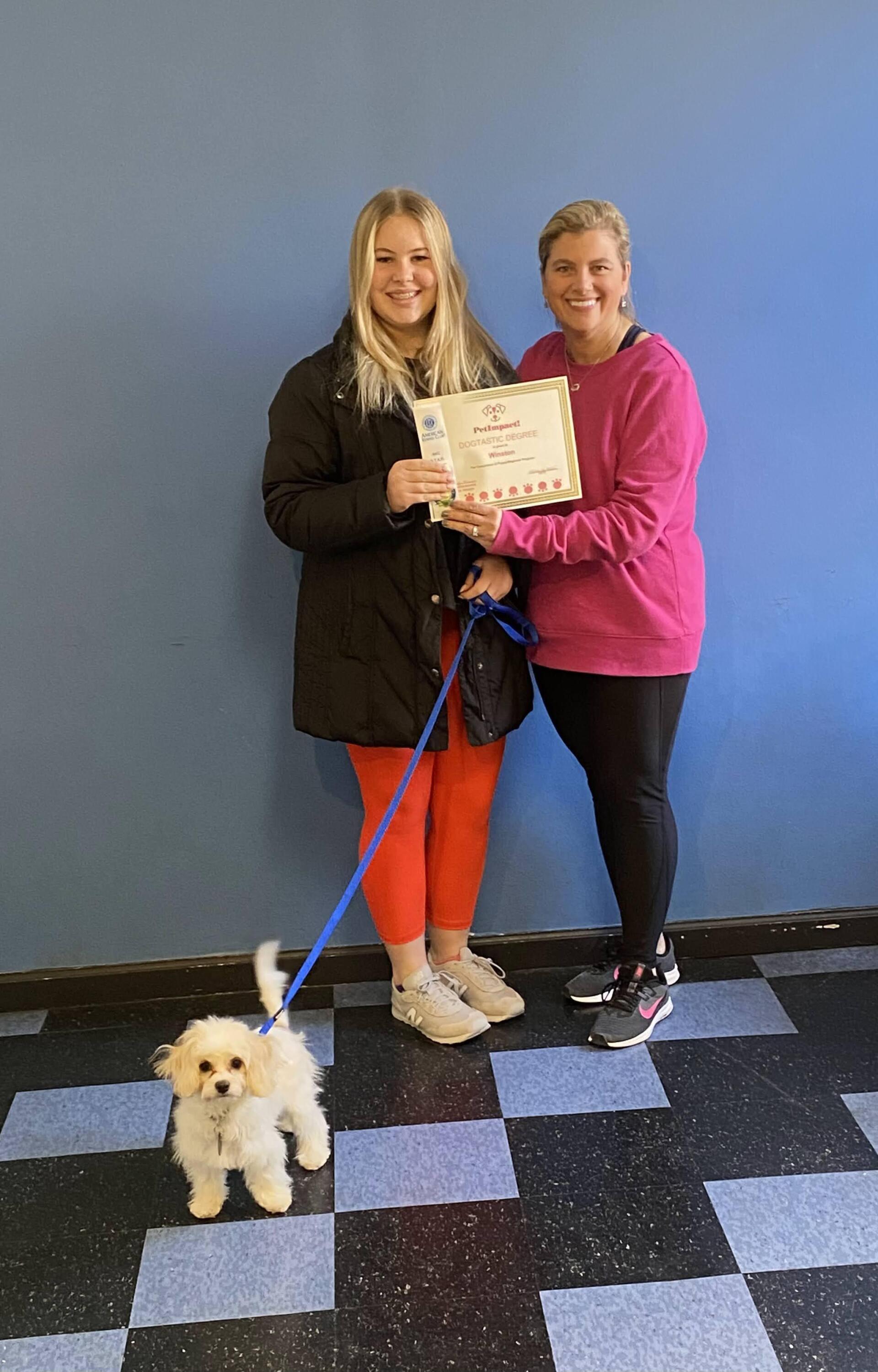 Woman holding certificate with another woman and dog. Blue wall.