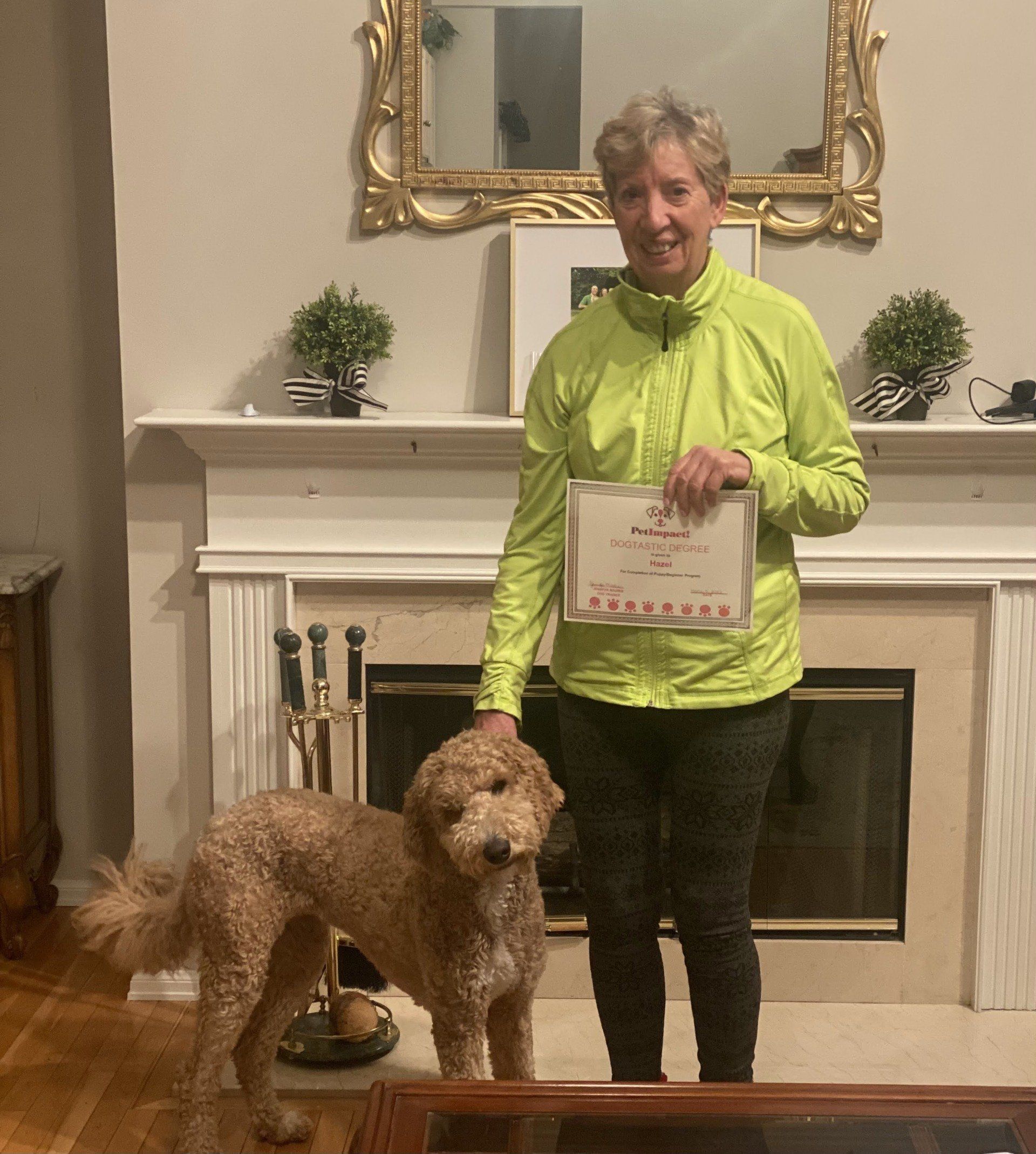 Woman in neon jacket and dog pose by fireplace, holding a certificate.