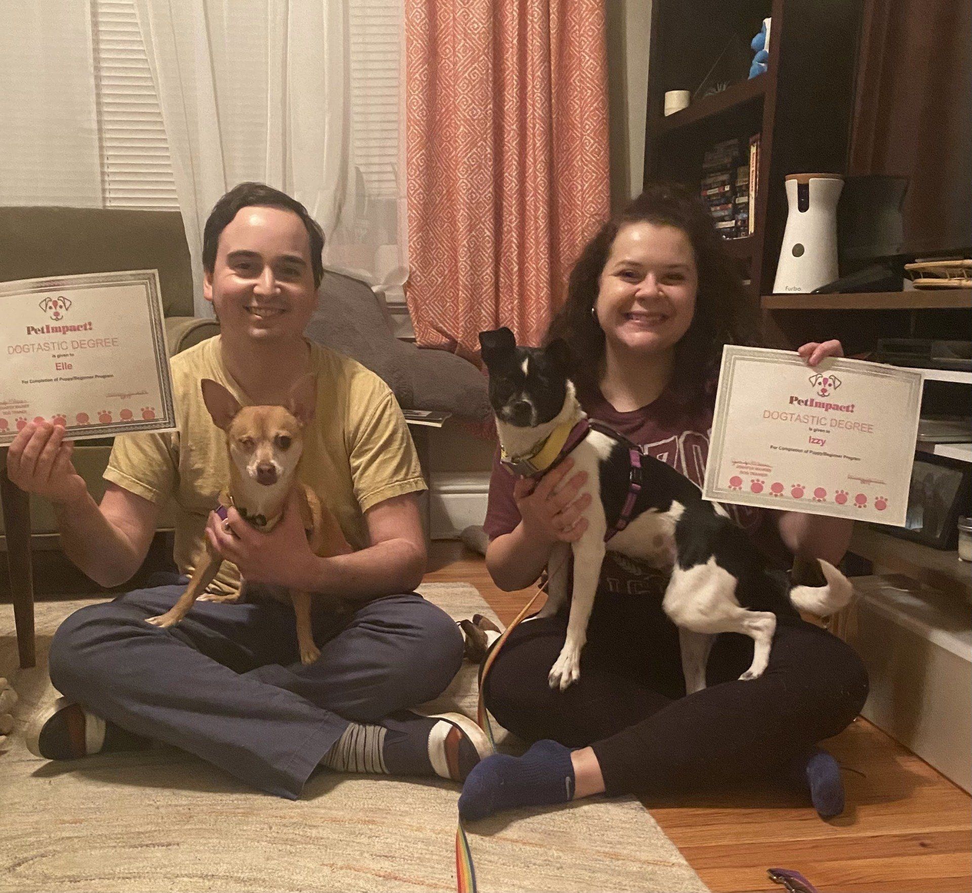 Two people sitting with their dogs, holding certificates. They are smiling. Indoor setting.