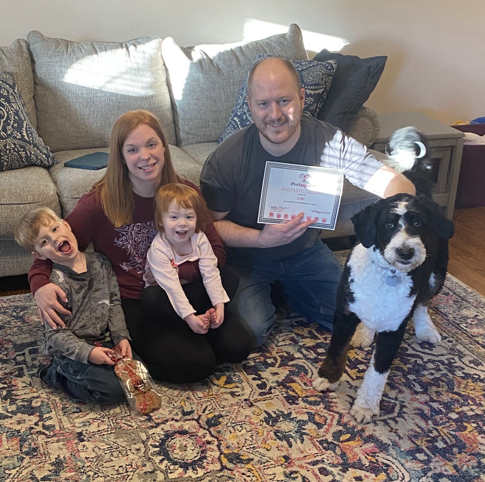 Family and dog on rug; man holds document. Two children, woman, and black and white dog. Sunlight.