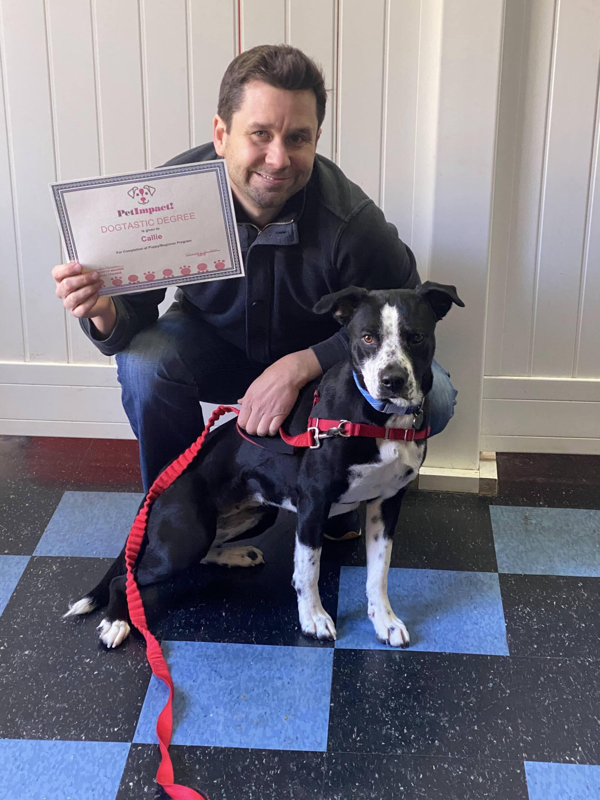 Man kneeling with a black and white dog, both smiling, holding adoption certificate. Blue and black tiled floor.